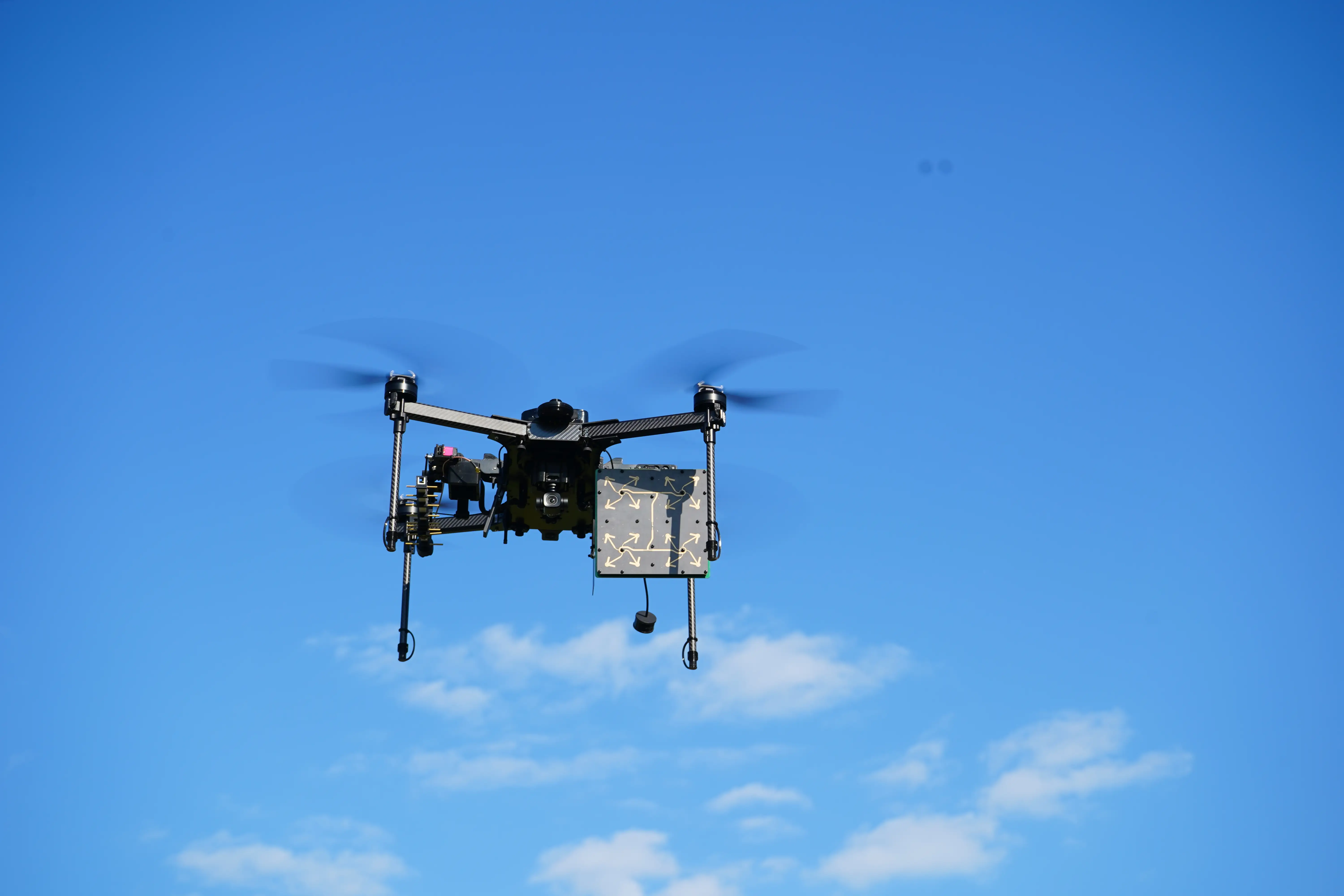 Quadcopter drone carrying a mounted payload flies against a bright blue sky with scattered clouds.
