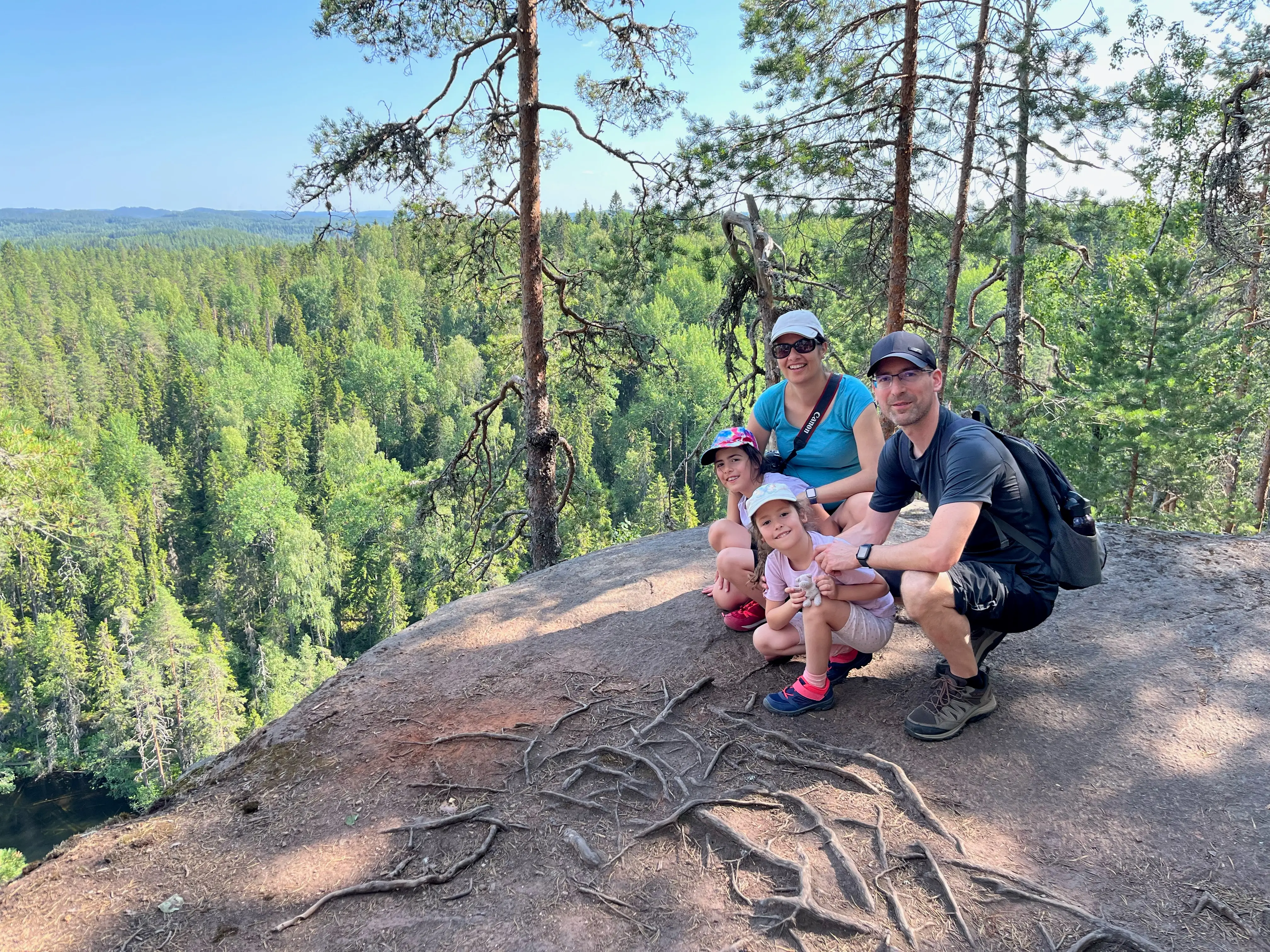Four hikers pose on a rocky forest overlook with expansive green woodland and distant hills behind them at Konnevesi National Park in Finland.