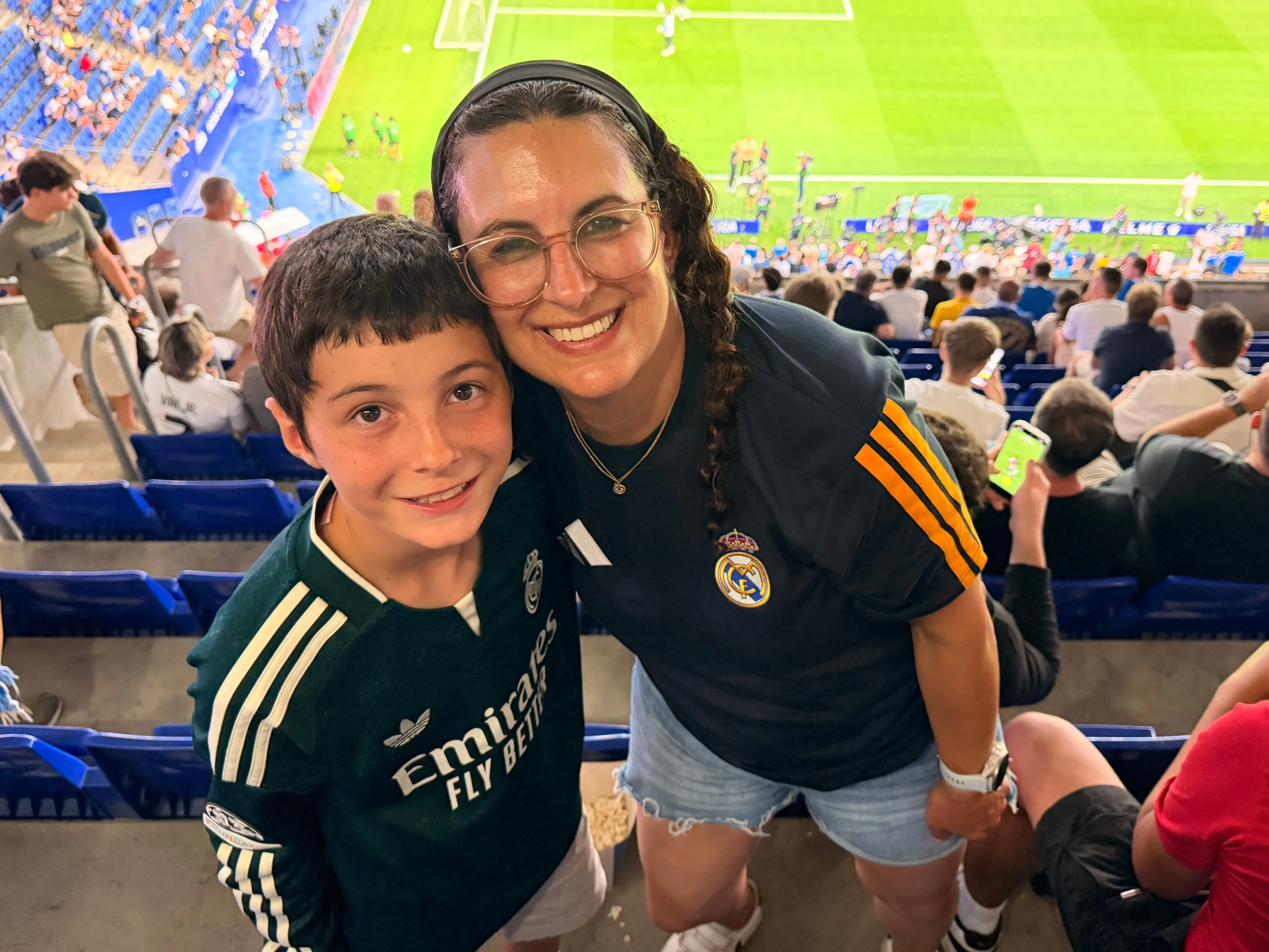Two Real Madrid supporters smile from stadium seats overlooking a soccer field during a match.