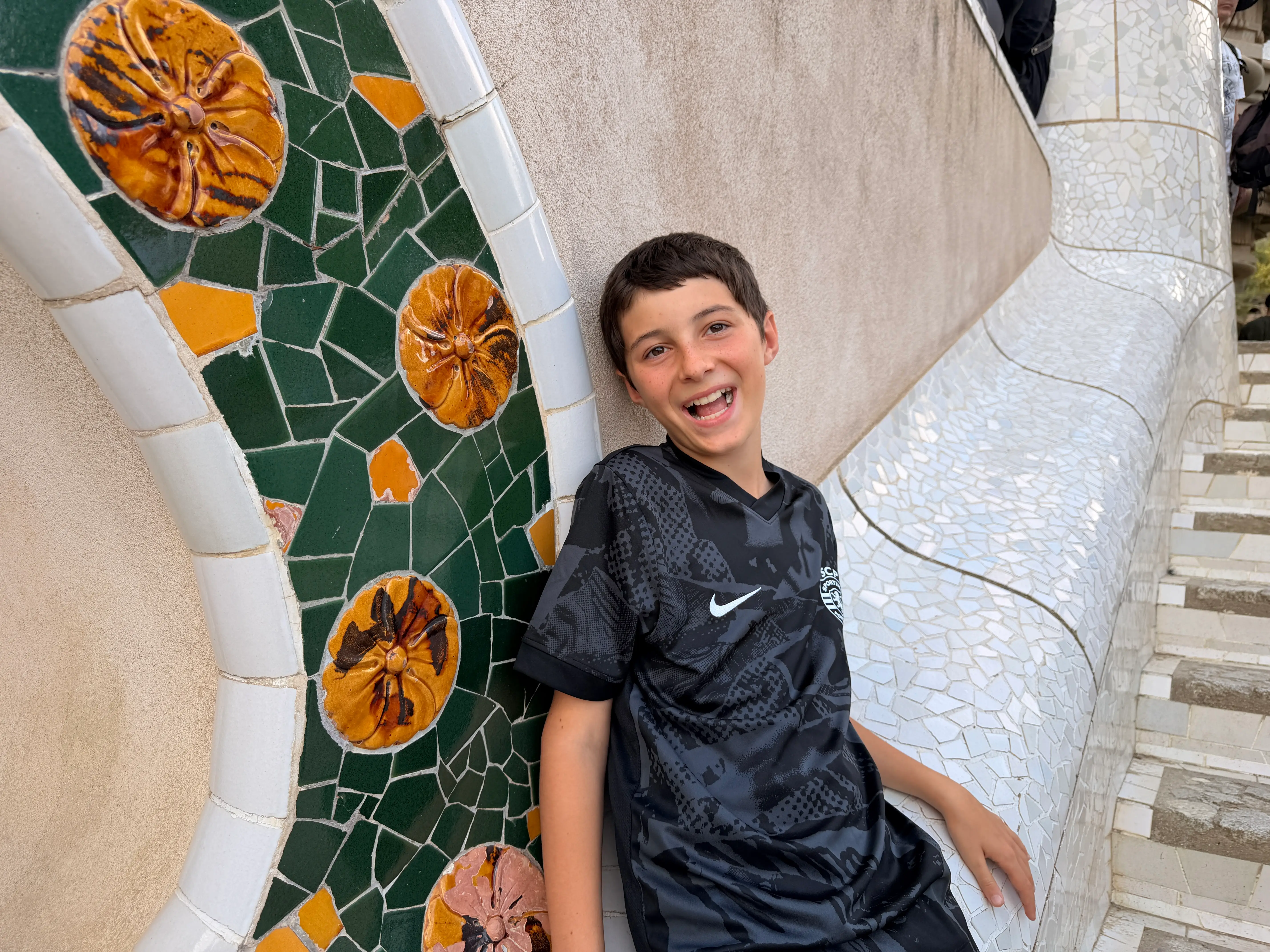 Smiling person in a black Nike soccer jersey sits beside colorful mosaic tiles at Park Güell.