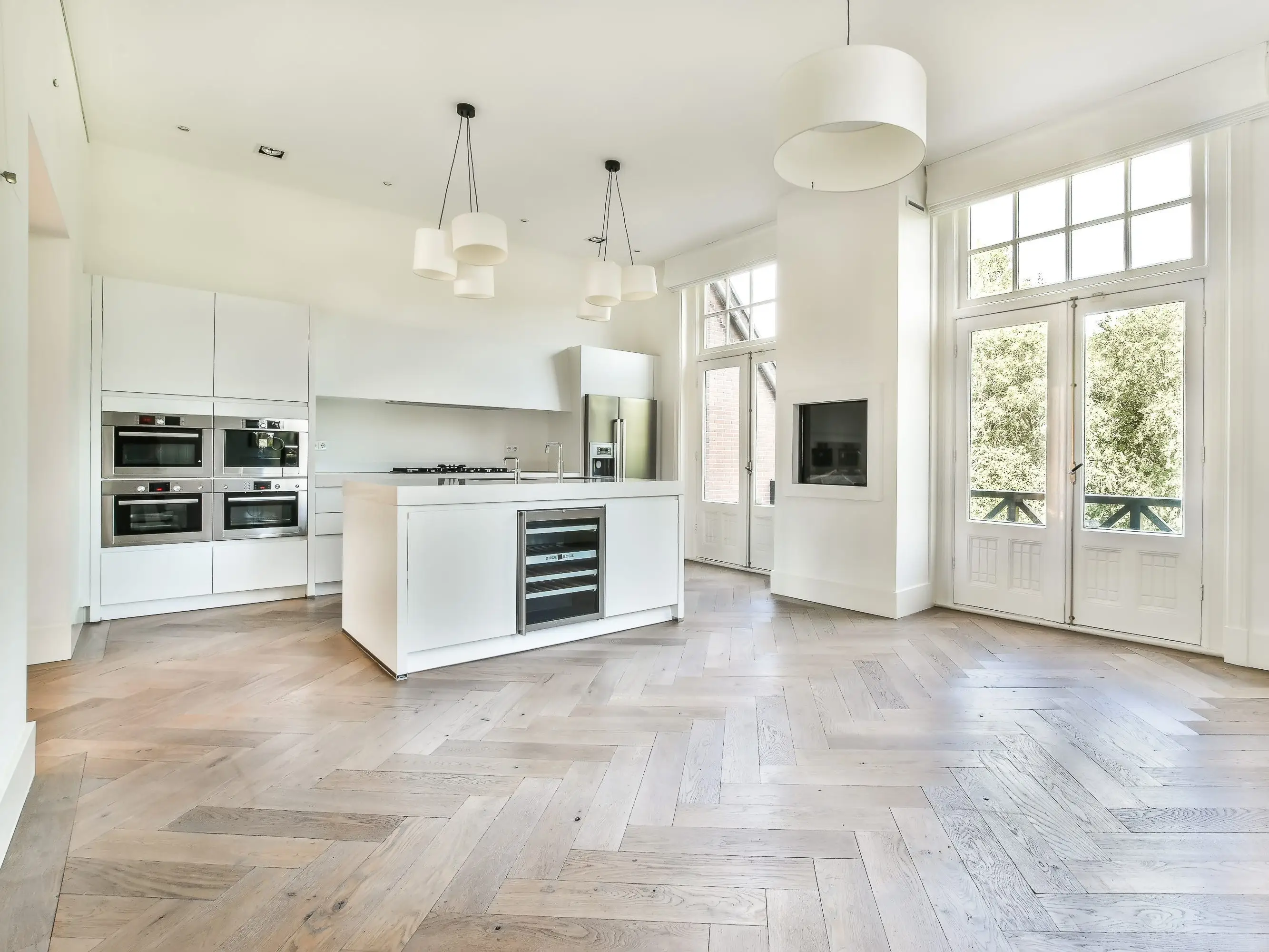 Bright open-plan kitchen with white cabinetry, island appliances, pendant lights, tall windows, and herringbone wood flooring.