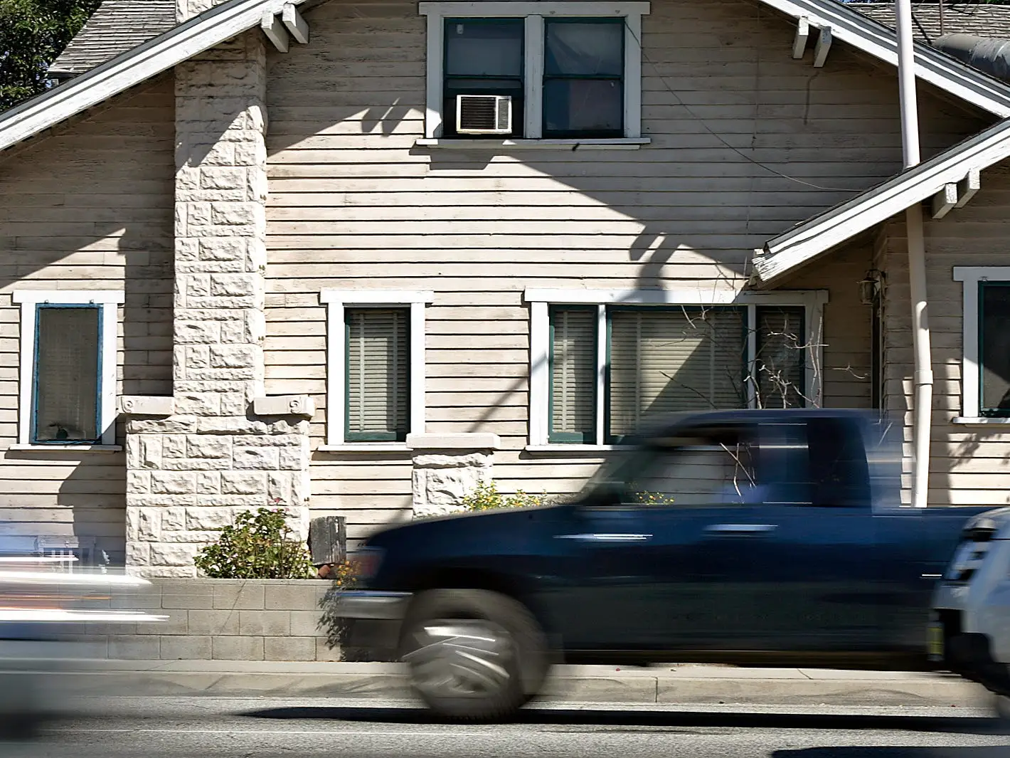 Blurred traffic passes in front of a beige wood-sided house with stone pillars on a sunny street.