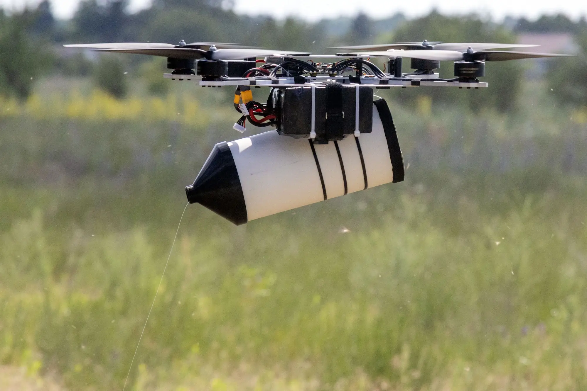 Multirotor drone flies low over a grassy field carrying a strapped cylindrical payload.