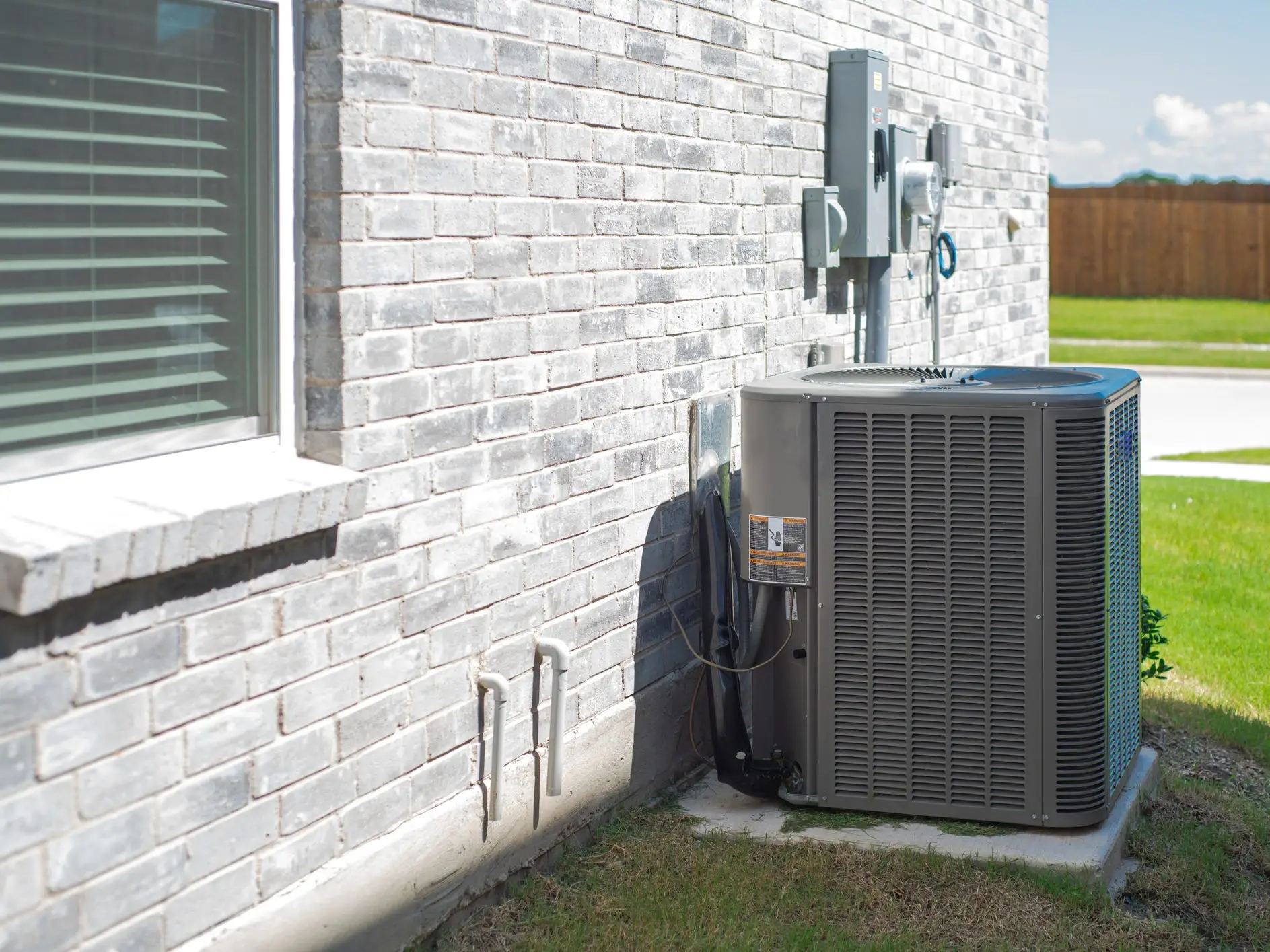 Outdoor HVAC condenser unit sits beside a light brick house wall with utility boxes, window, lawn, and fence.