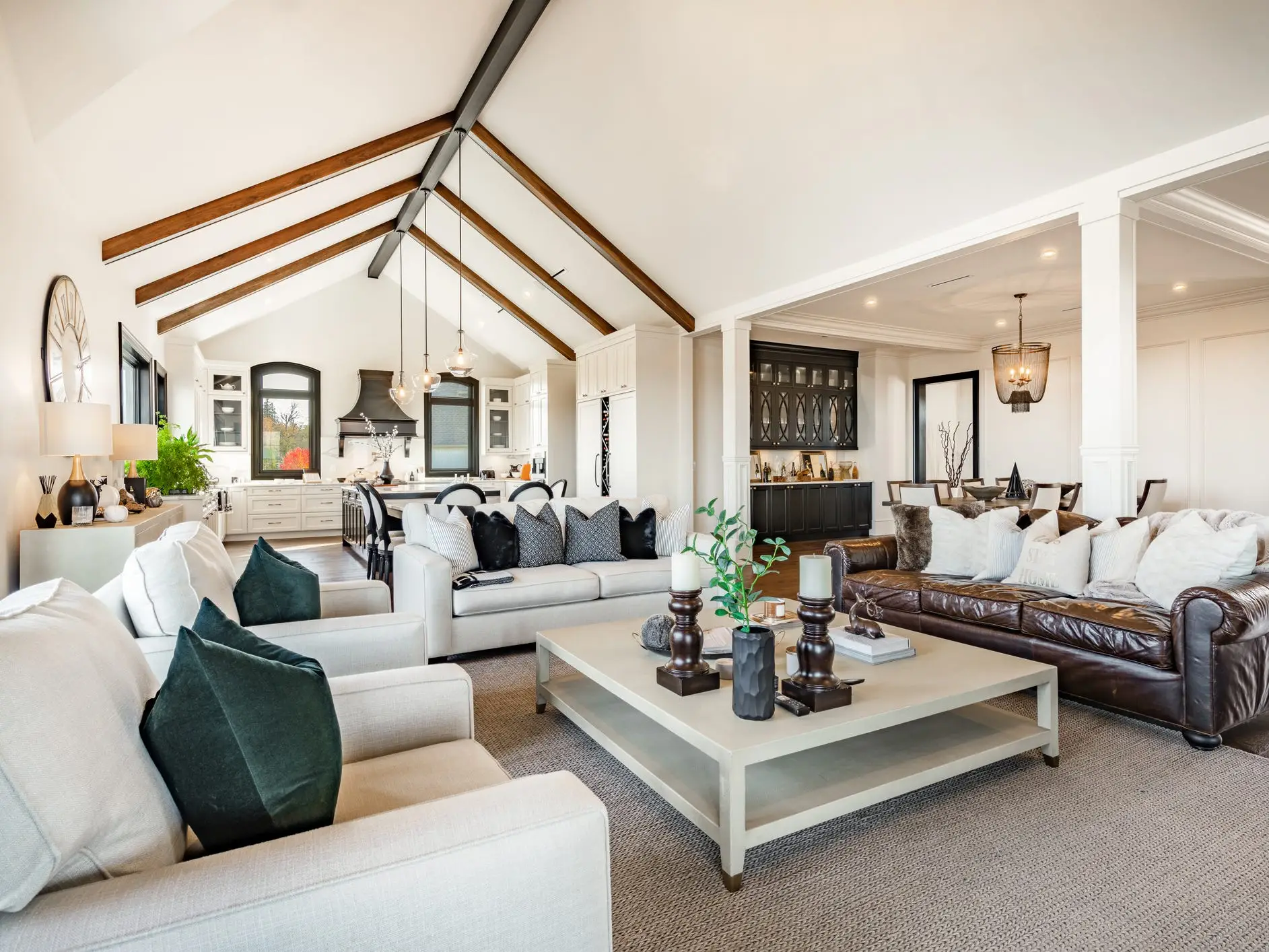 Open-concept living room with sofas, coffee table, vaulted ceiling beams, kitchen, and dining area in bright neutral decor.