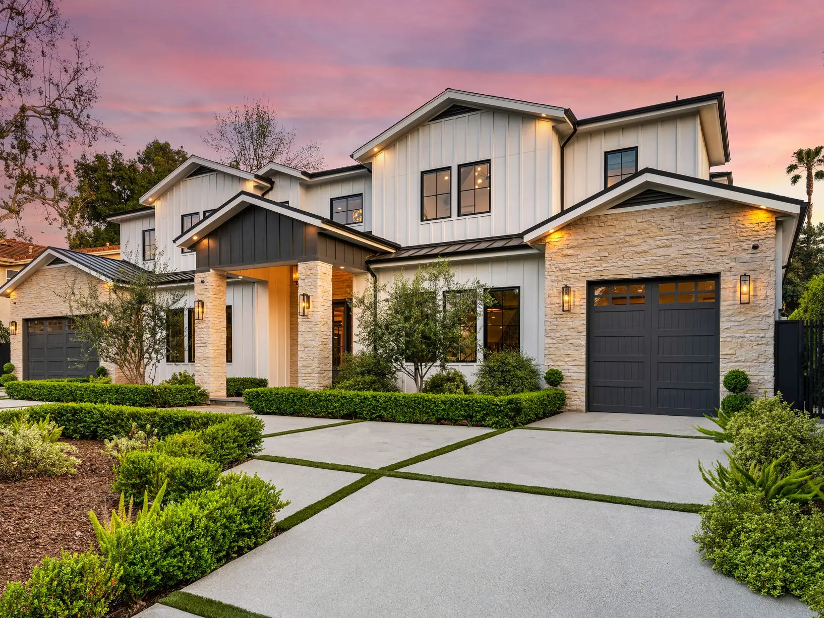 Modern two-story house with landscaped front yard, garages, and exterior lights under a pink sunset sky.