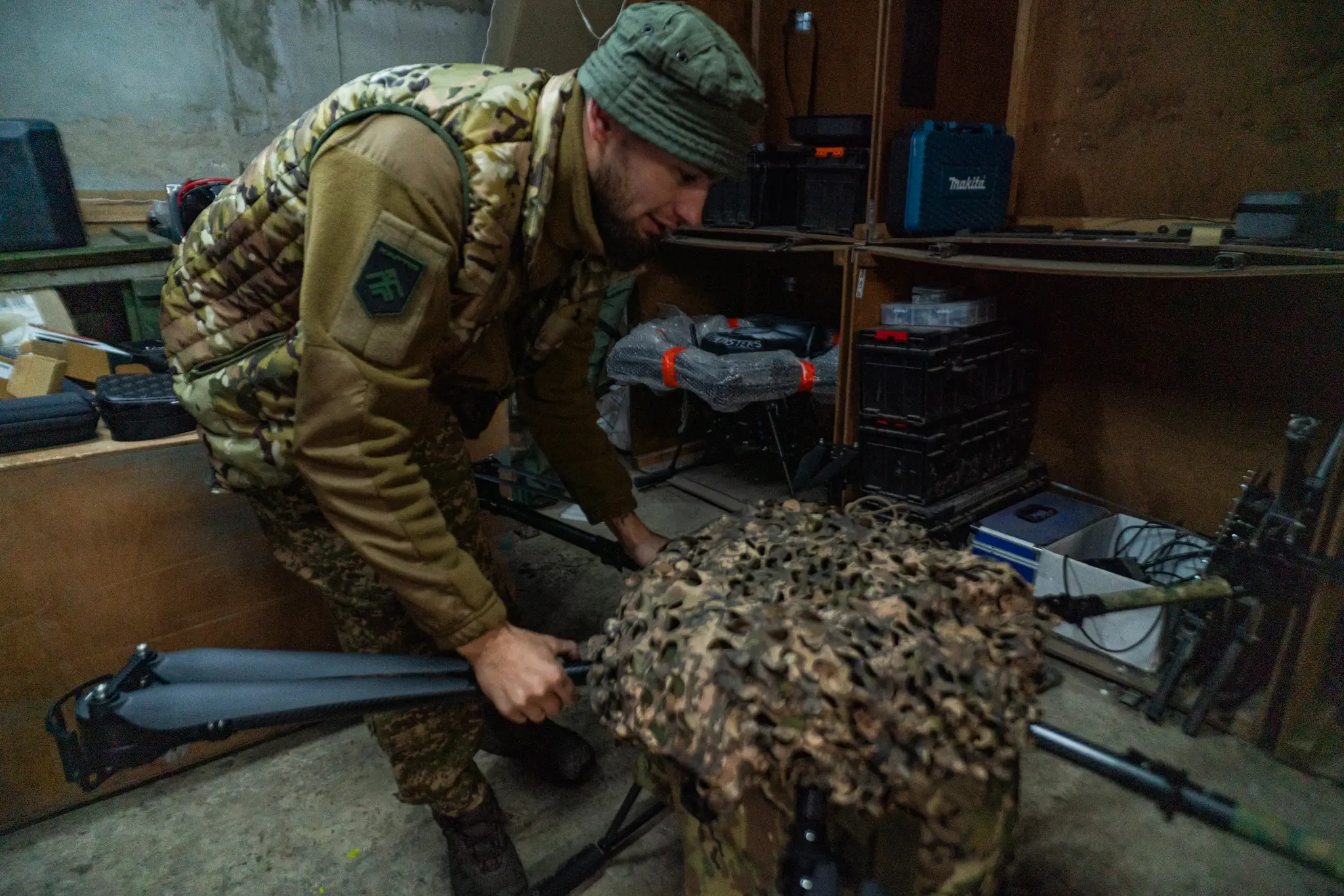 Camouflage-uniformed person adjusts a net-covered drone inside a workshop with tools and equipment.