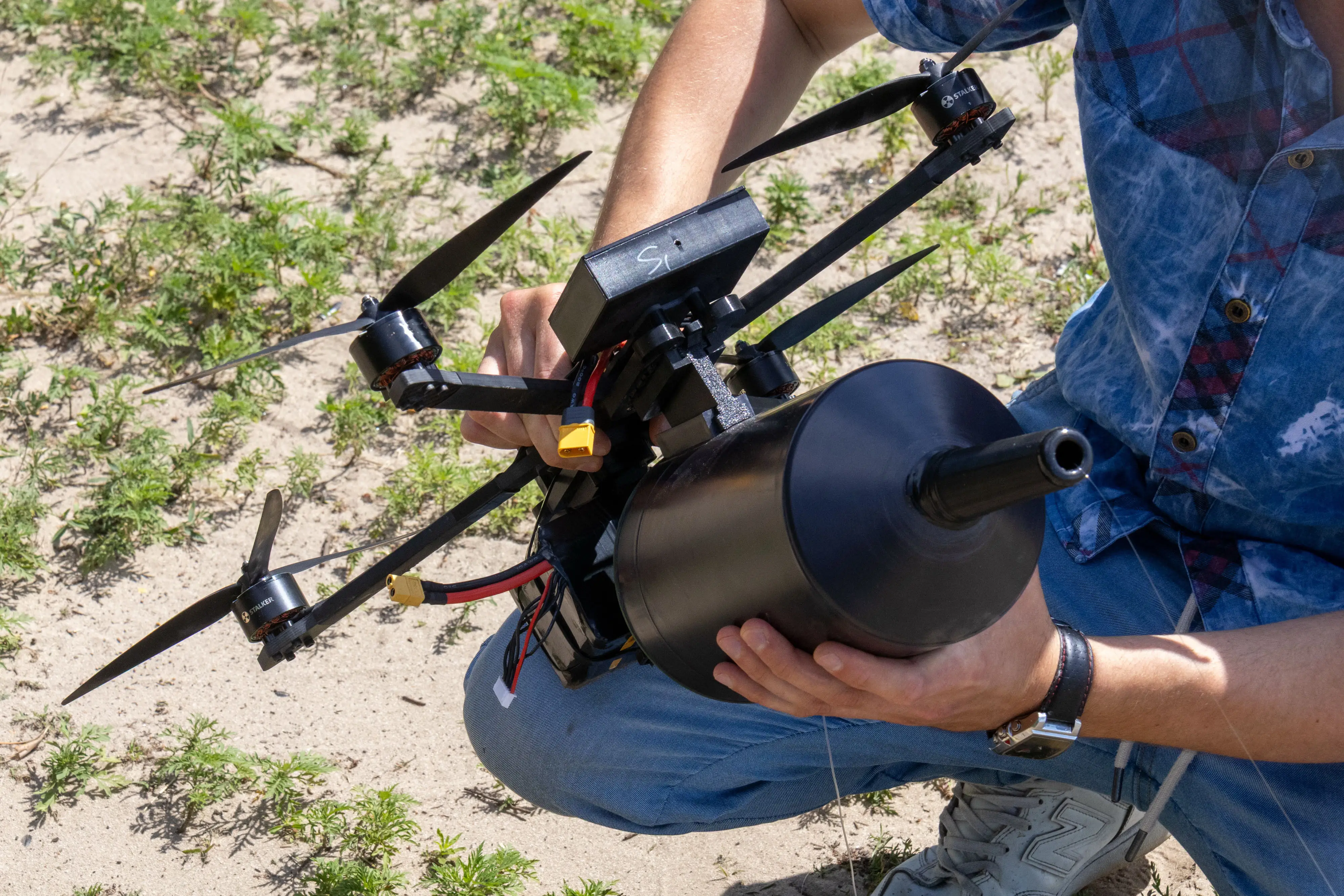 Unidentified person crouches outdoors while holding a quadcopter drone with a large black cylindrical attachment.