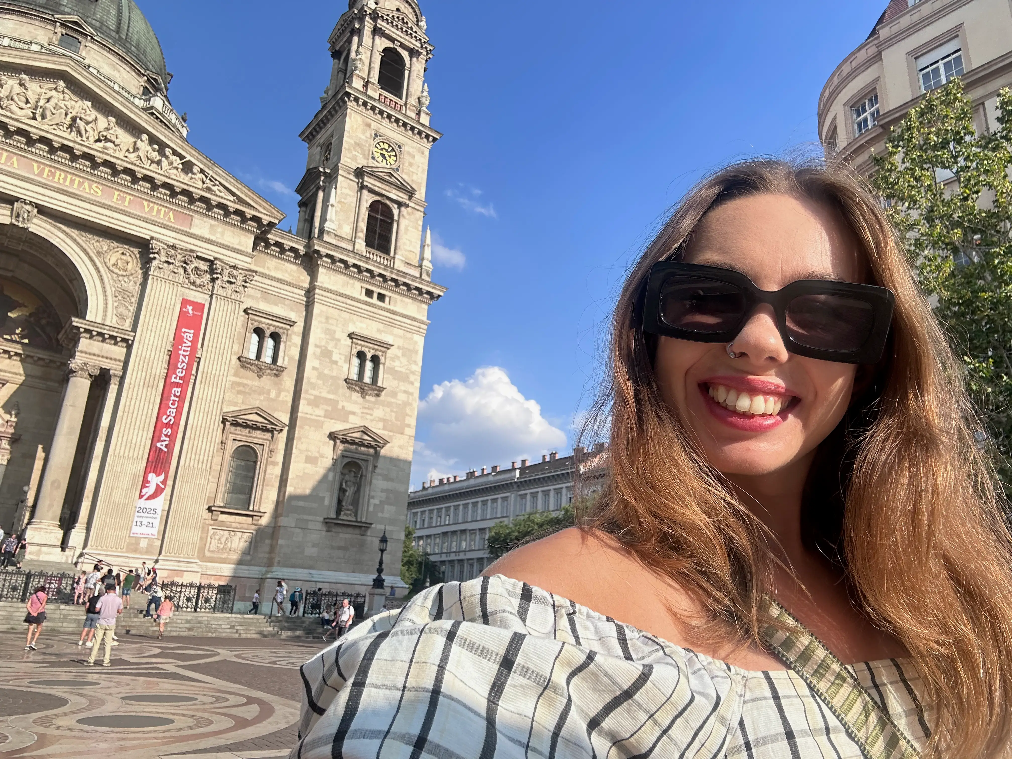 The writer wears black sunglasses and poses outside St. Stephen's Basilica with an Ars Sacra Fesztivál banner.