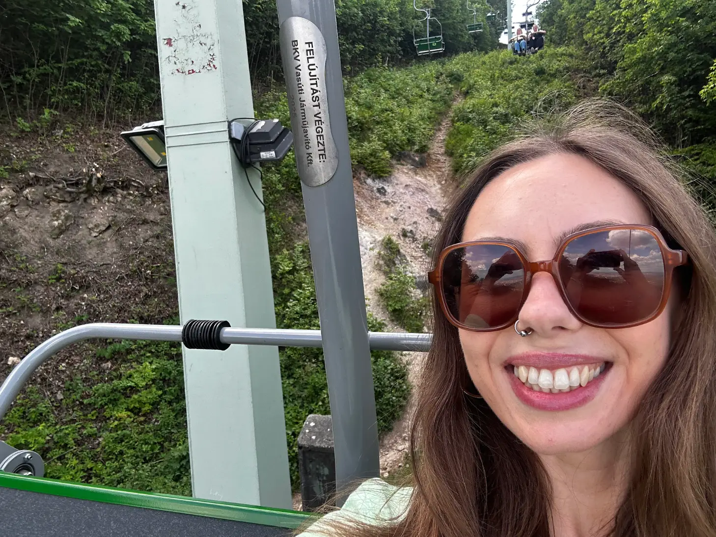 The writer smiles, wearing sunglasses and riding a chairlift above a green wooded hillside.