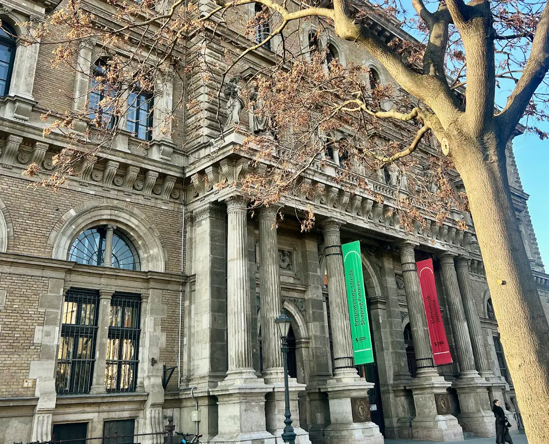 Ornate stone-and-brick building facade with columns, statues, banners, and bare tree branches in front.