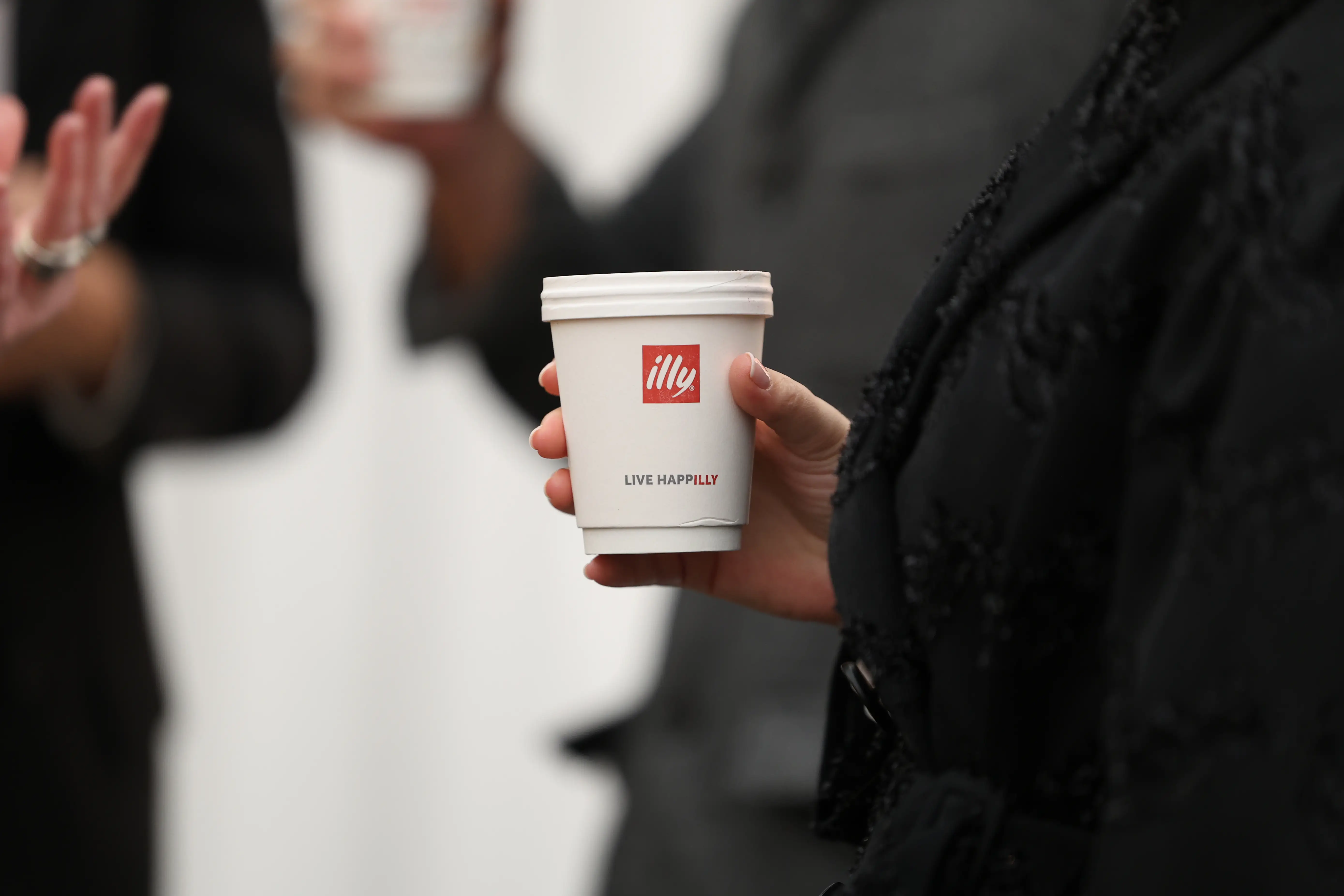 A person in dark clothing holds a white illy paper coffee cup at an indoor gathering.