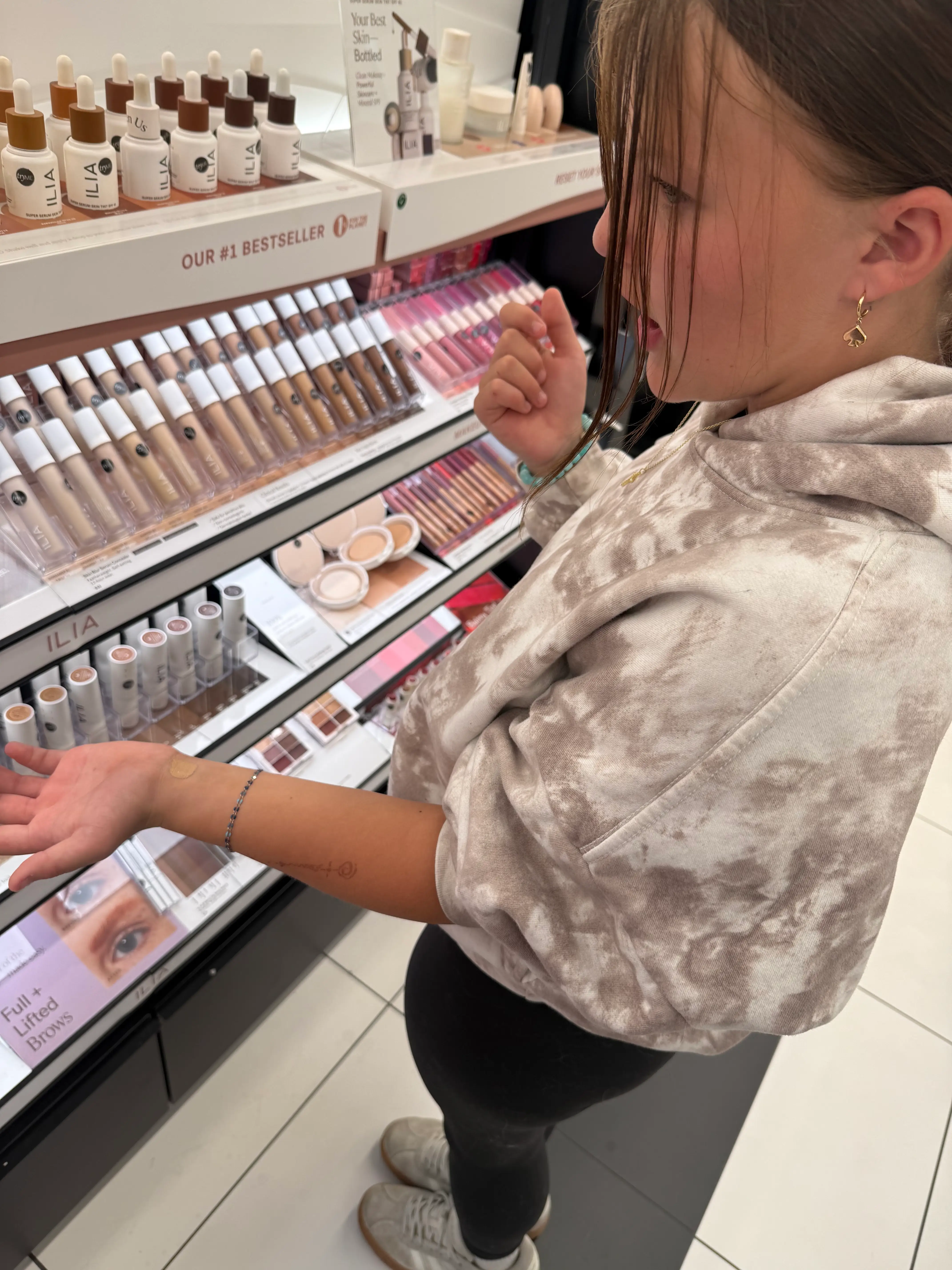 Shopper tests makeup on their hand beside an ILIA cosmetics display with foundation, lip, and brow products.