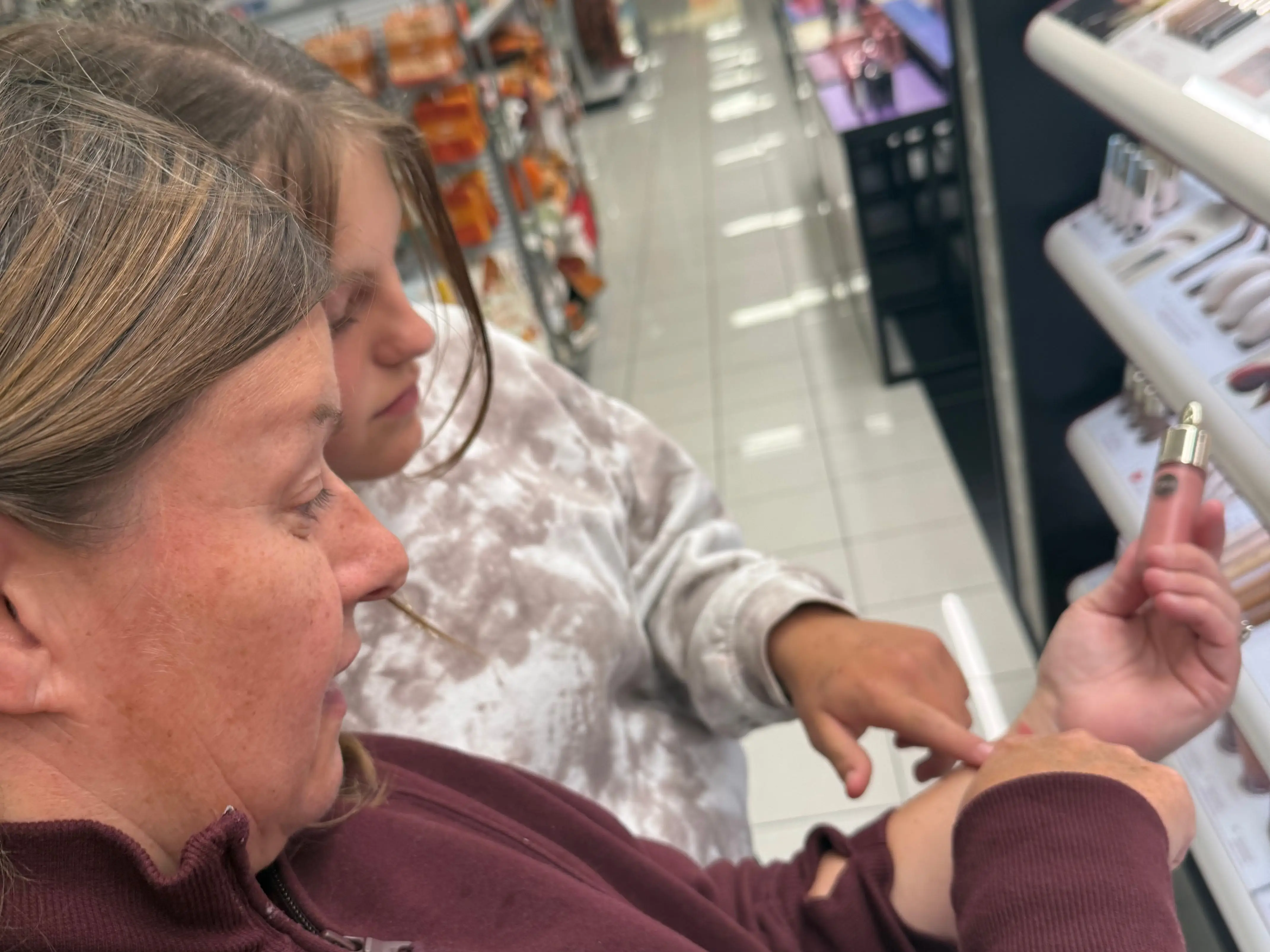 Two shoppers examine a small pink cosmetic product beside makeup displays in a brightly lit retail store.