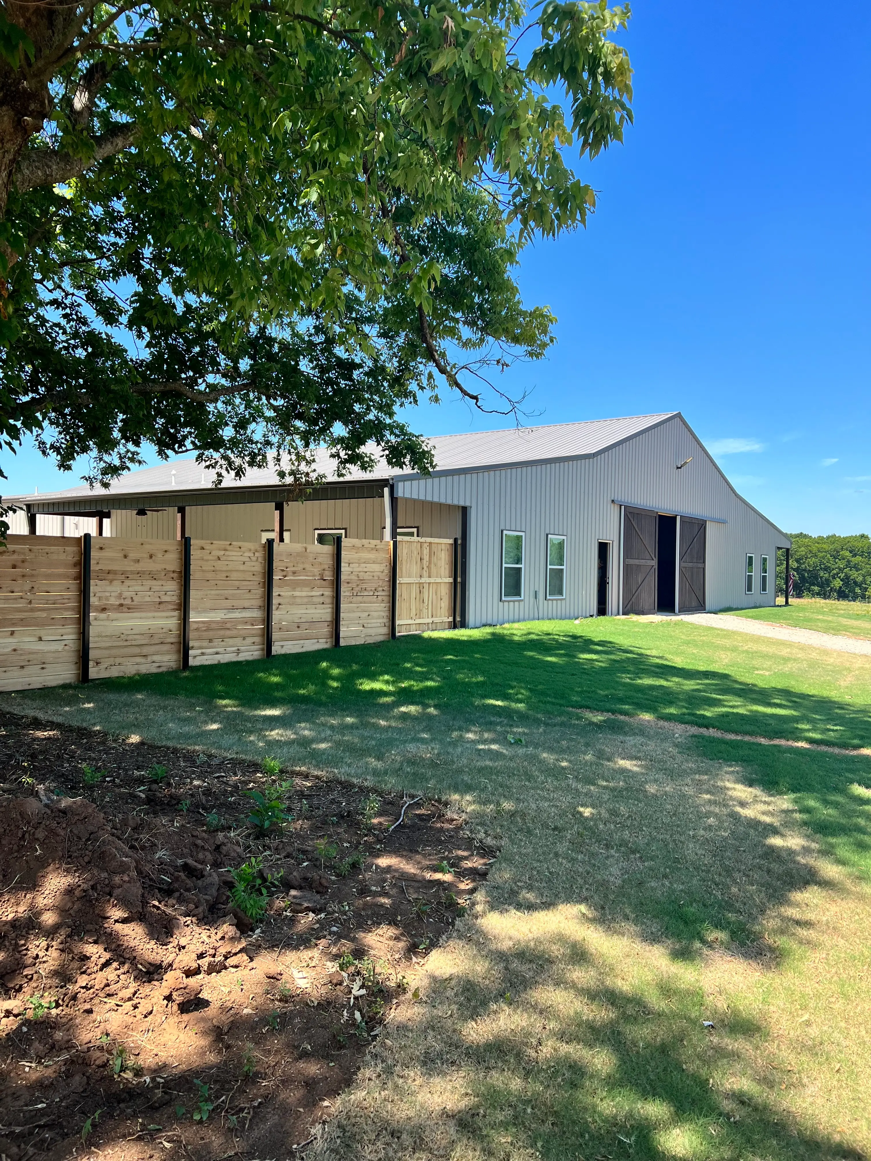 Metal barn with sliding doors stands beside a wooden fence under a leafy tree and blue sky.