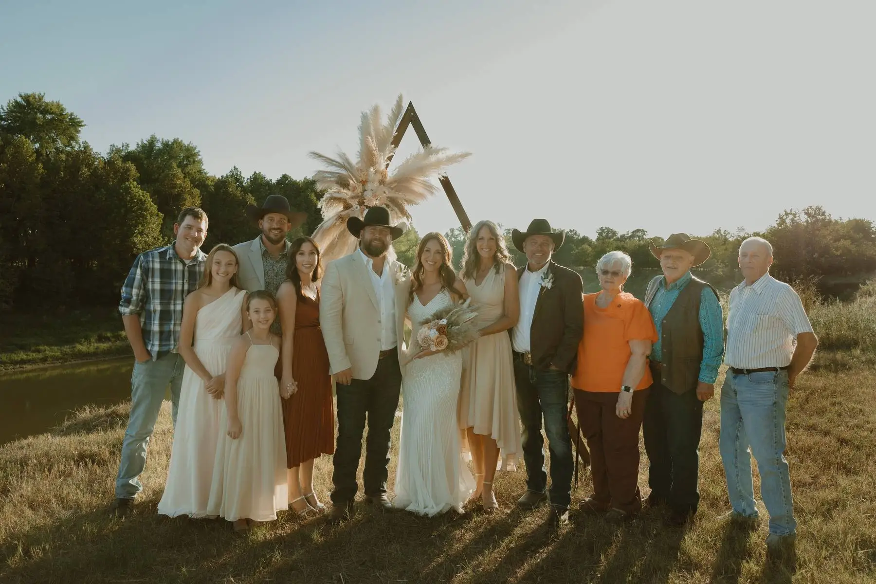 Large wedding group poses outdoors near a pond with bouquets, cowboy hats, and a floral arch.