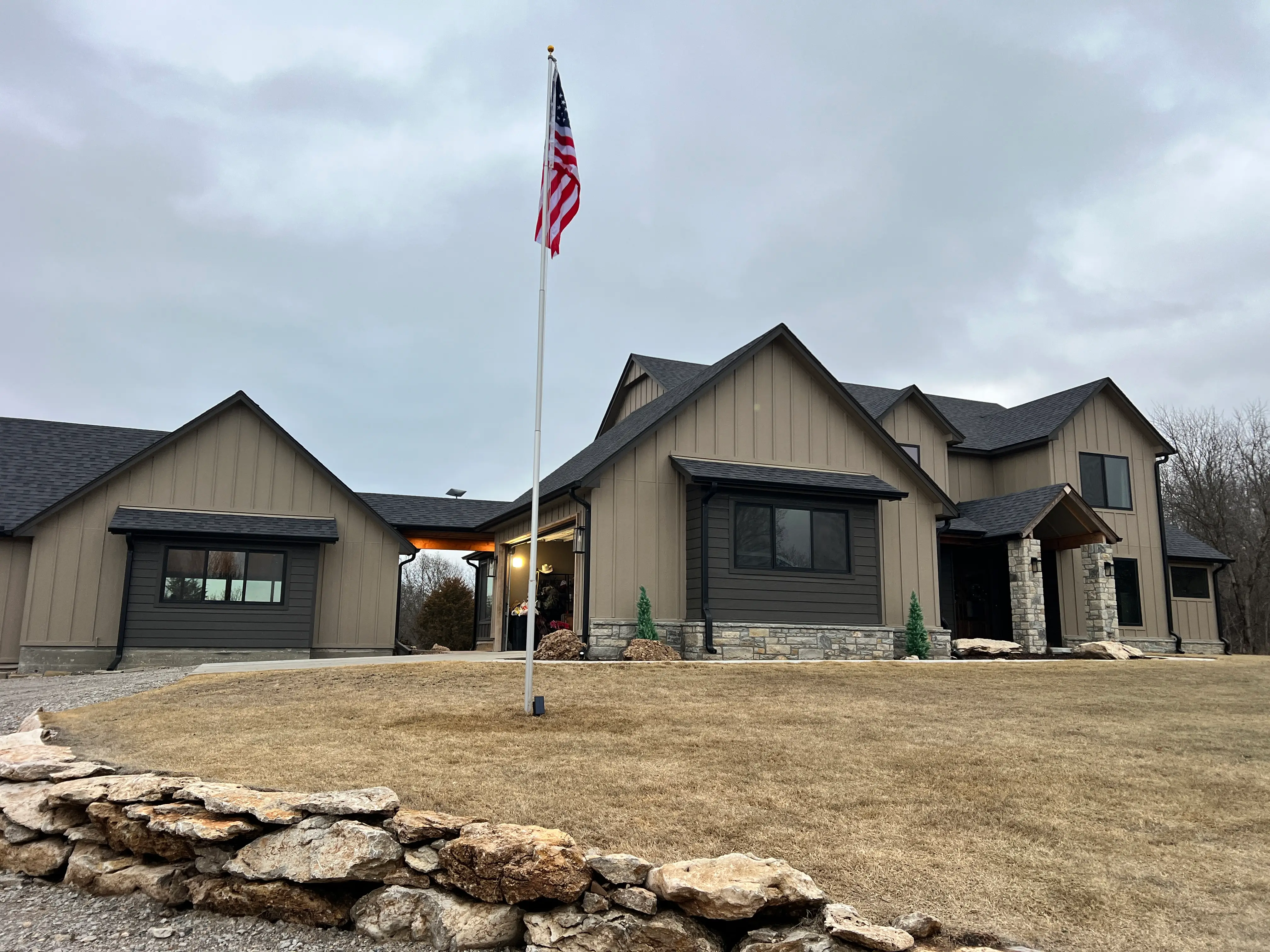 Modern farmhouse-style home with an American flag on the front lawn under an overcast sky.