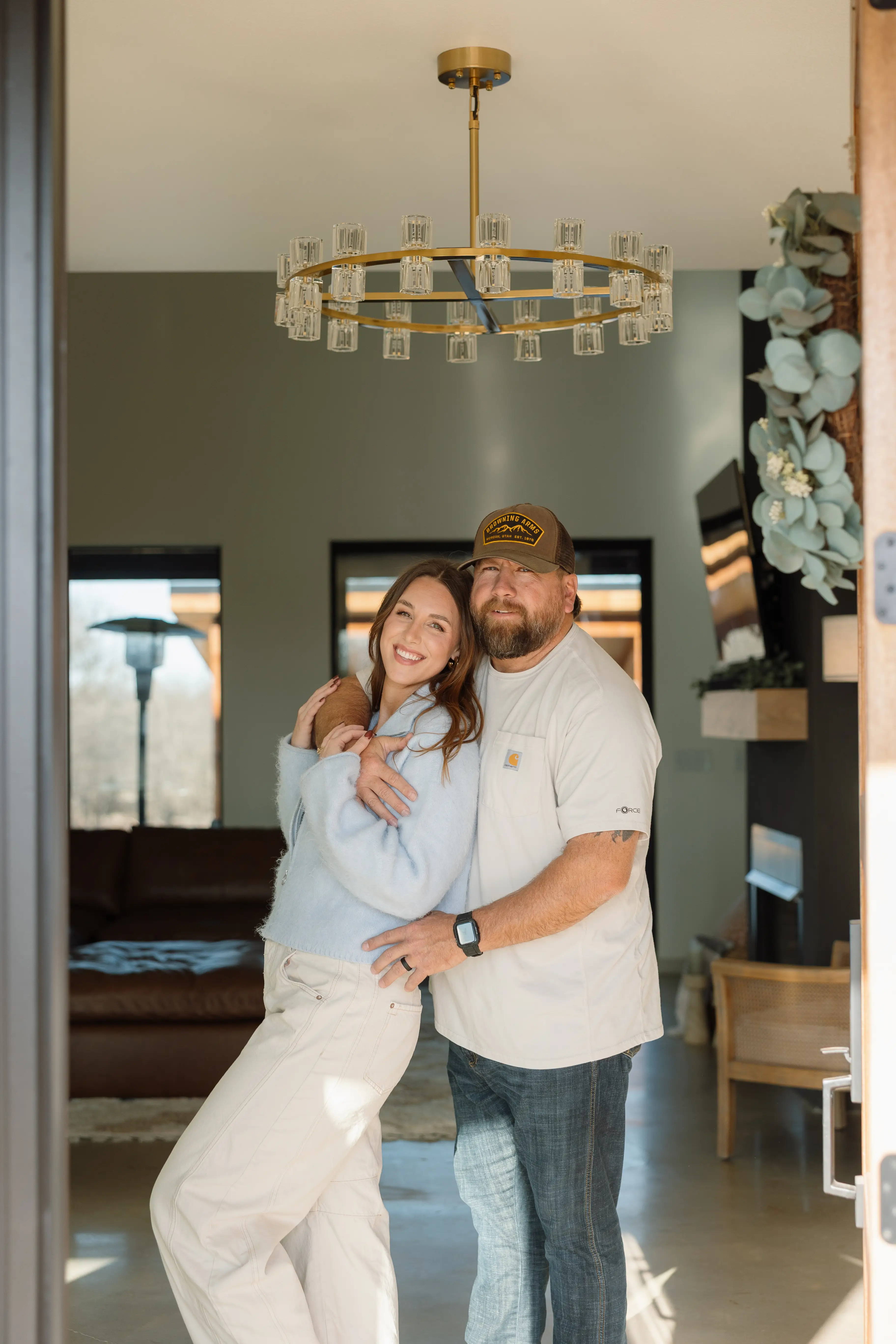 Two people smile and embrace in a sunlit home entryway beneath a circular chandelier.