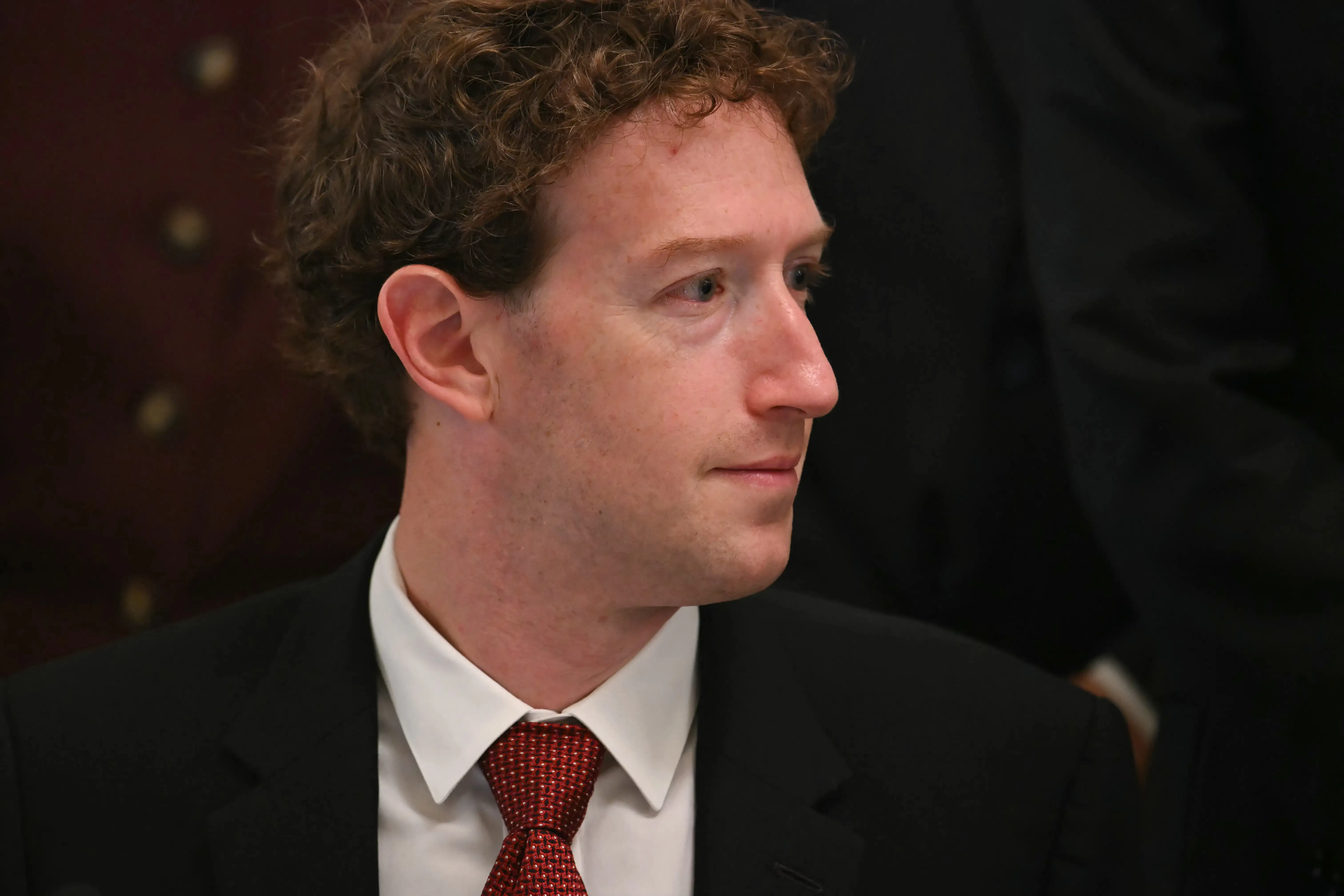Mark Zuckerberg sits in a black suit and red tie, looking to the side in an indoor formal setting.