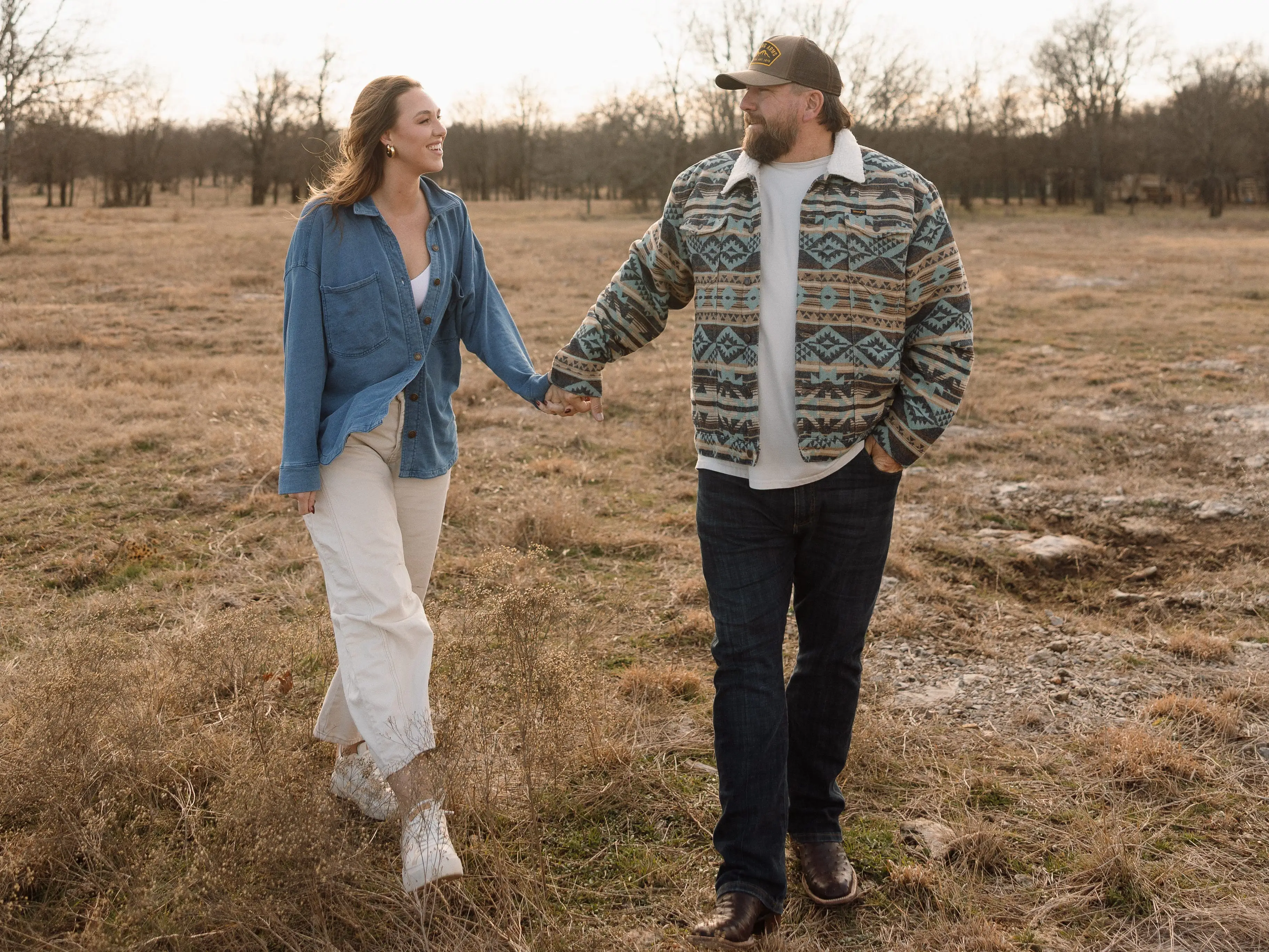 Two people hold hands while walking through a dry grassy field with bare trees in the background.