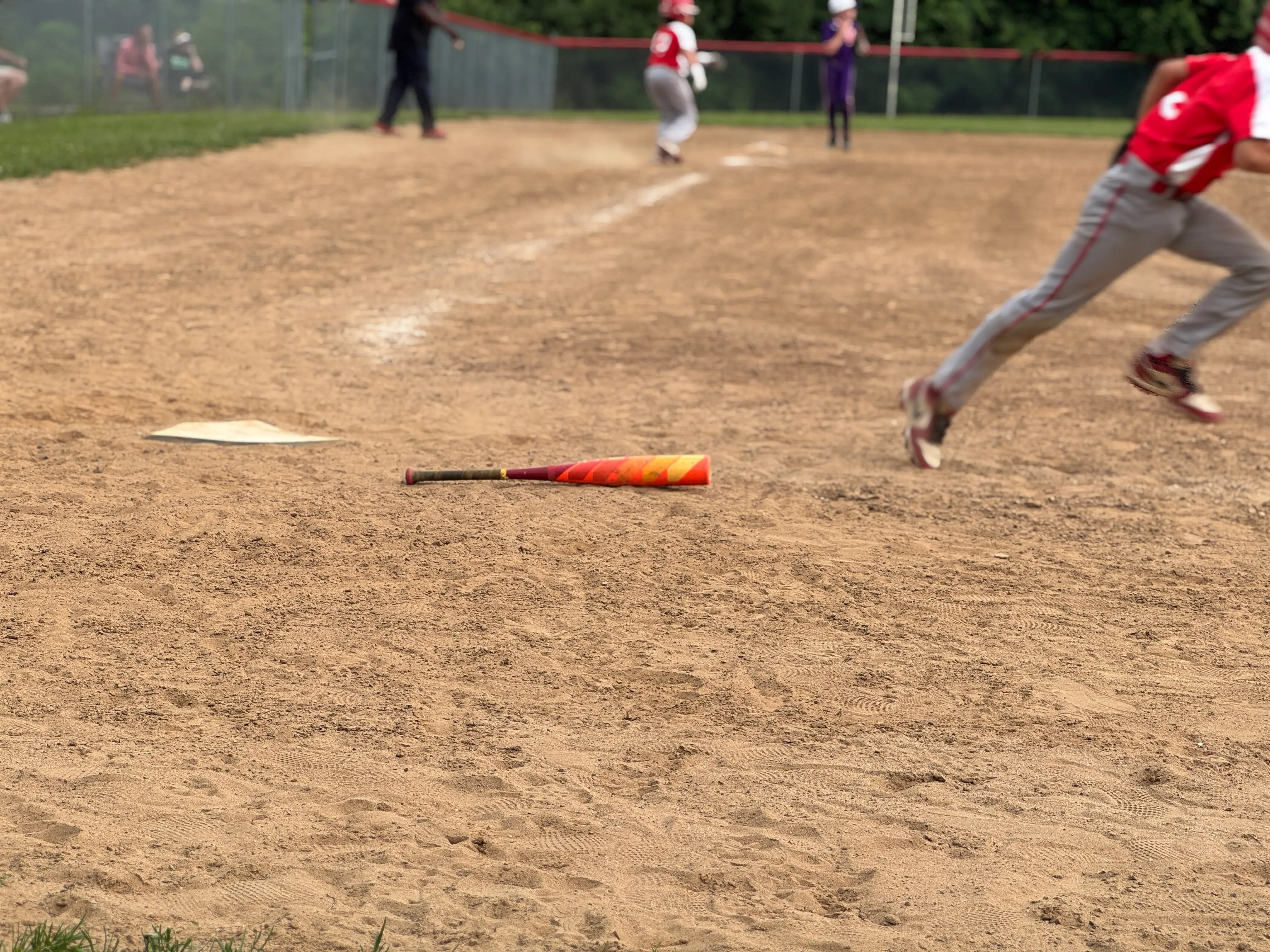 A baseball bat lies on a dusty infield as players run during a youth baseball game.