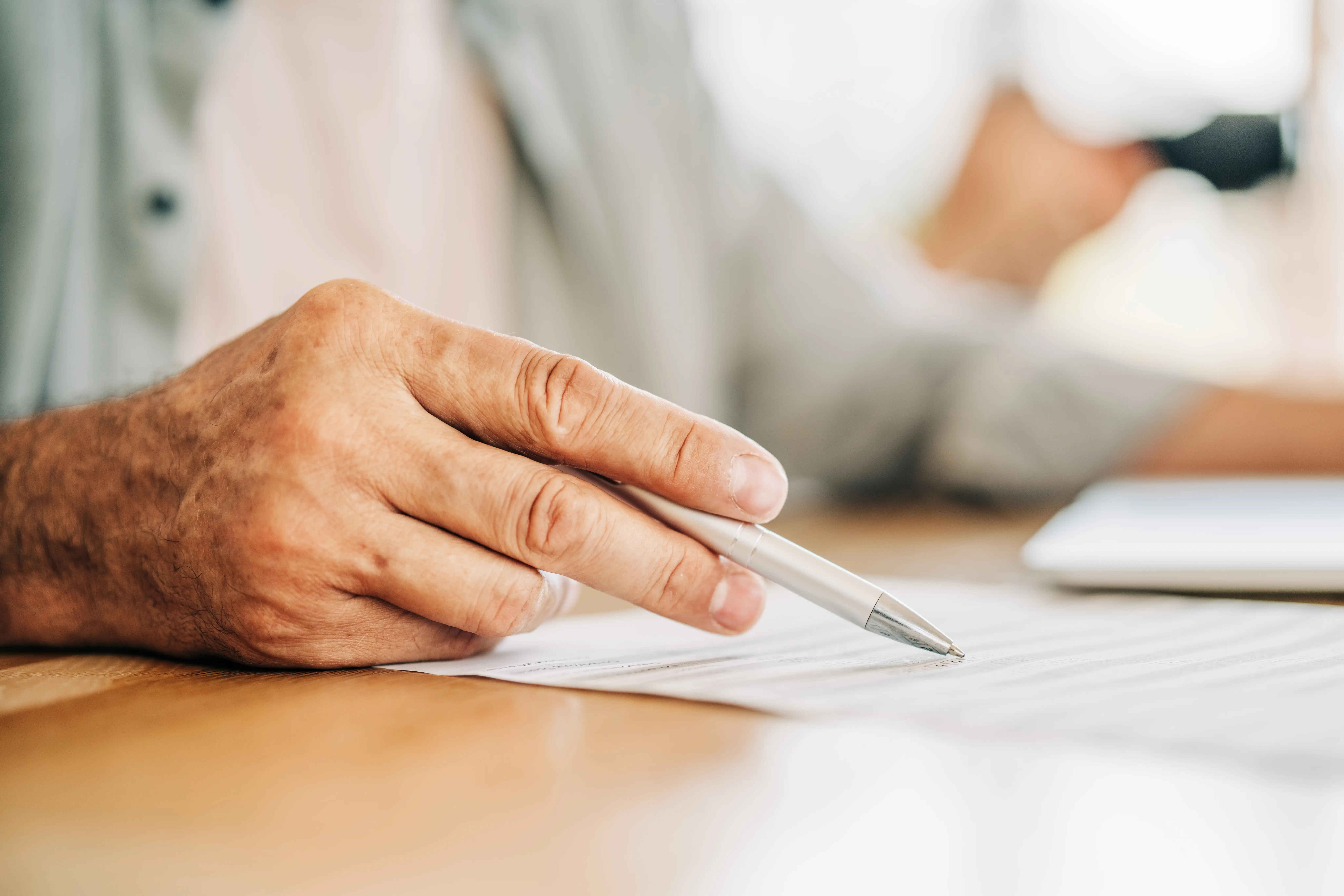 A close-up shot of an older person's hand holding a pen, pointing at a document