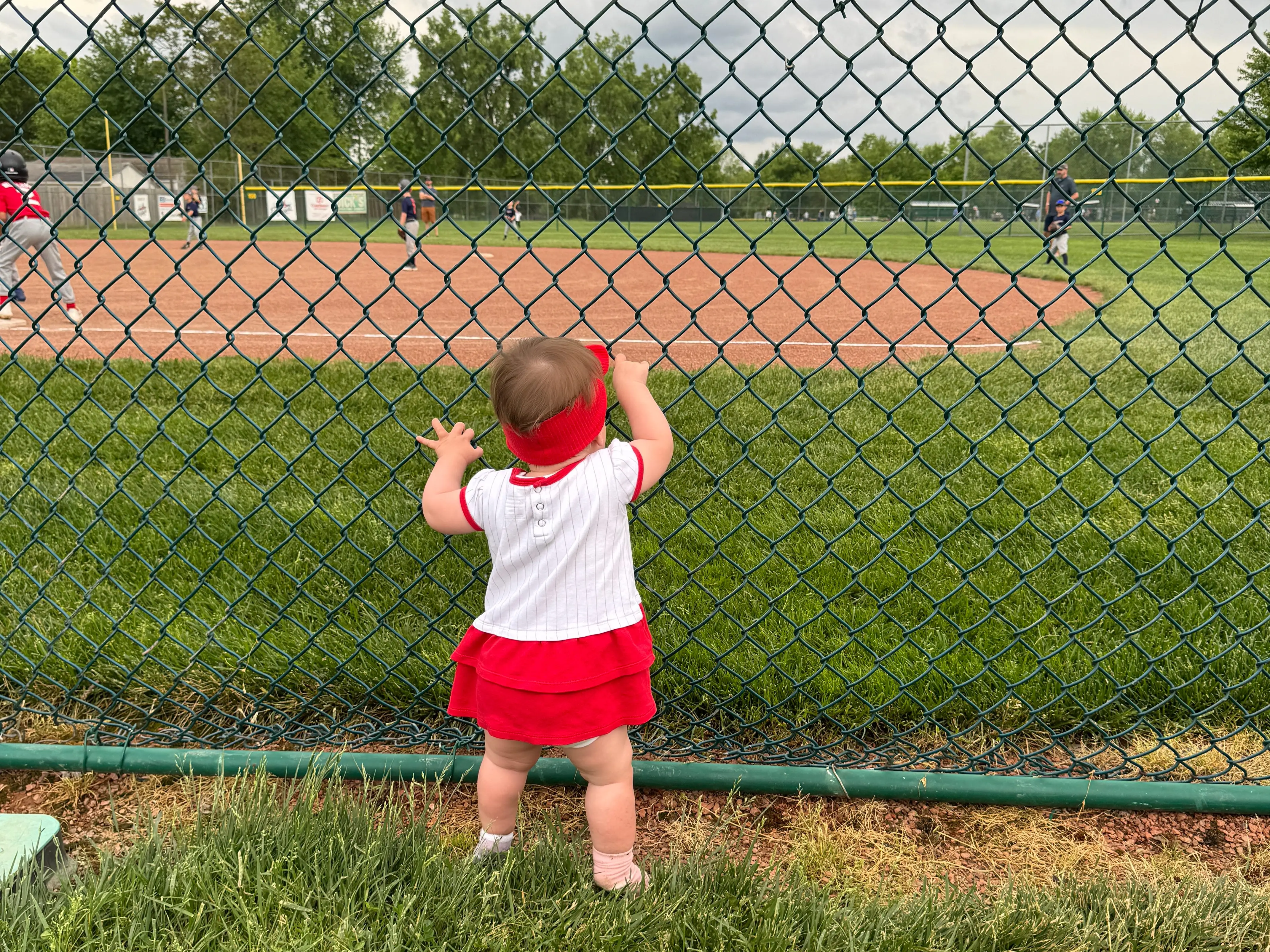 A spectator in red and white stands at a chain-link fence watching a baseball game on a grassy field.