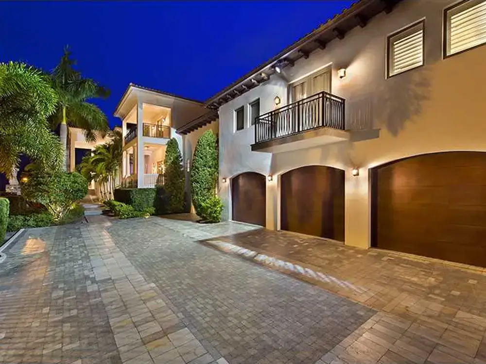 A Mediterranean-style residence with a balcony, three garage doors, and palm trees in the background.