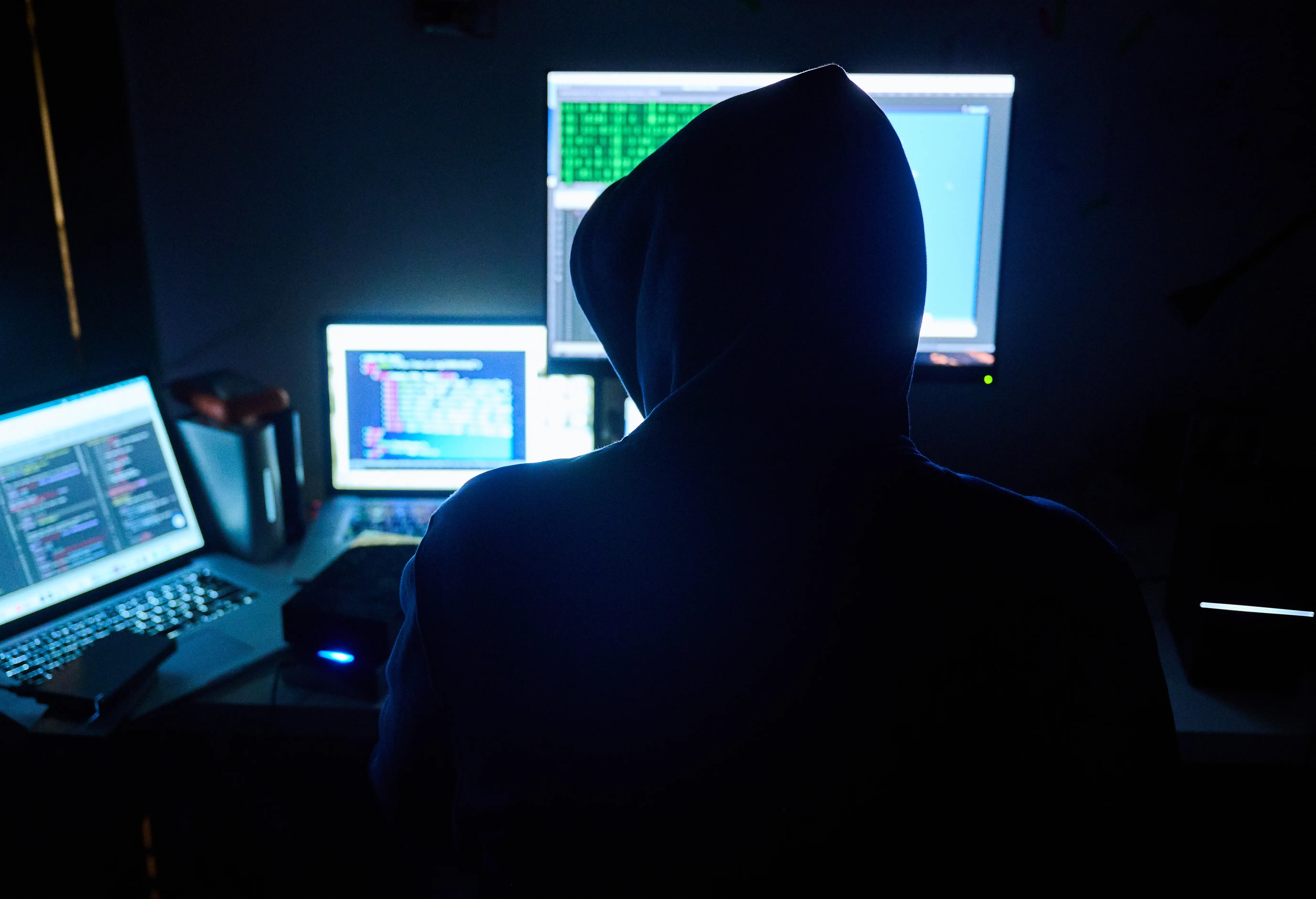 Hooded silhouette sits at a dark workstation facing multiple computer screens displaying code.