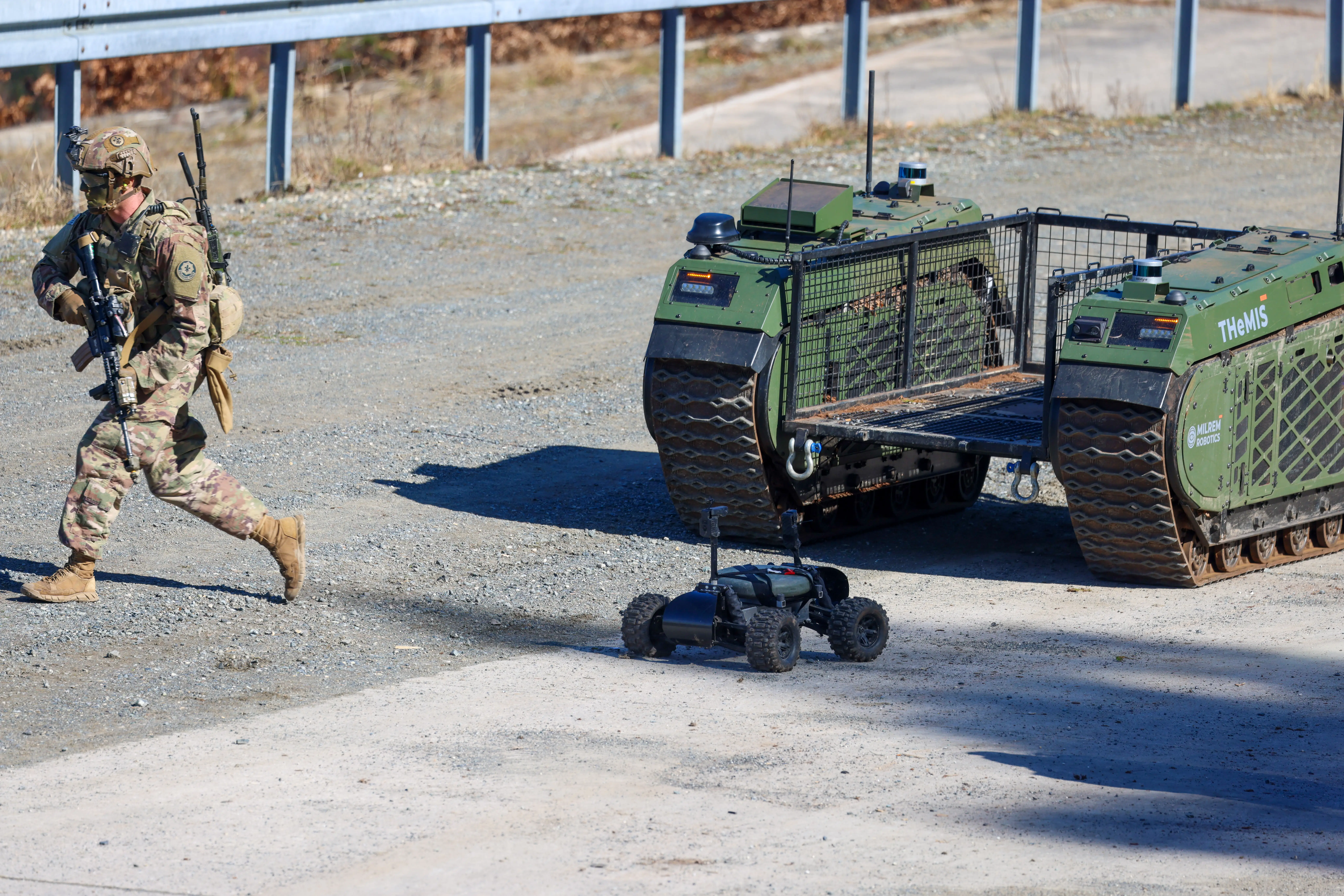 Camouflaged service member carrying a rifle walks beside THeMIS unmanned ground vehicles on a gravel training area.