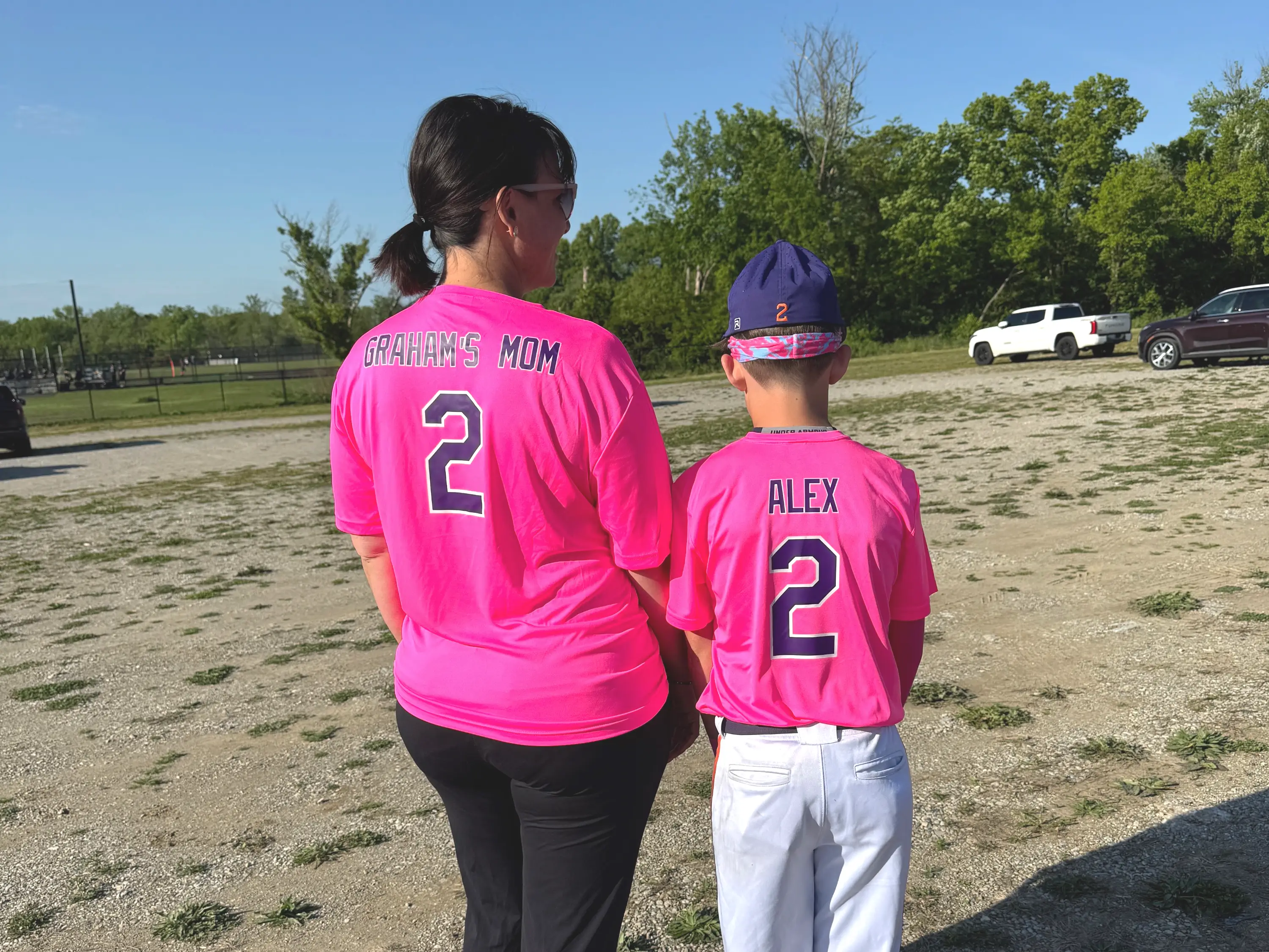 Two people stand with backs turned wearing bright pink baseball jerseys on a gravel field near parked cars.