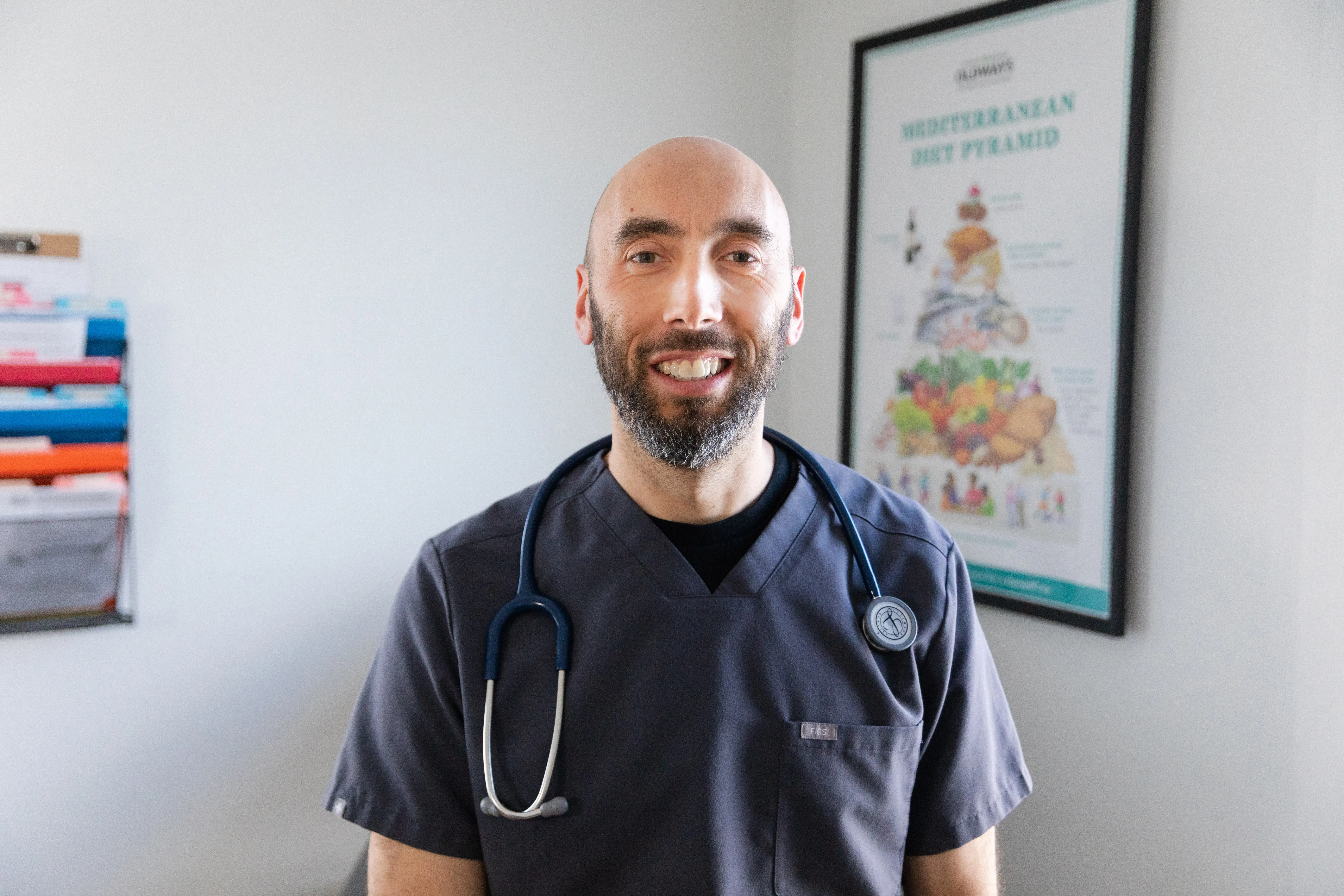 Smiling healthcare worker in dark scrubs with a stethoscope stands in a clinical office with a nutrition poster. Dr_Ben_Hagopian