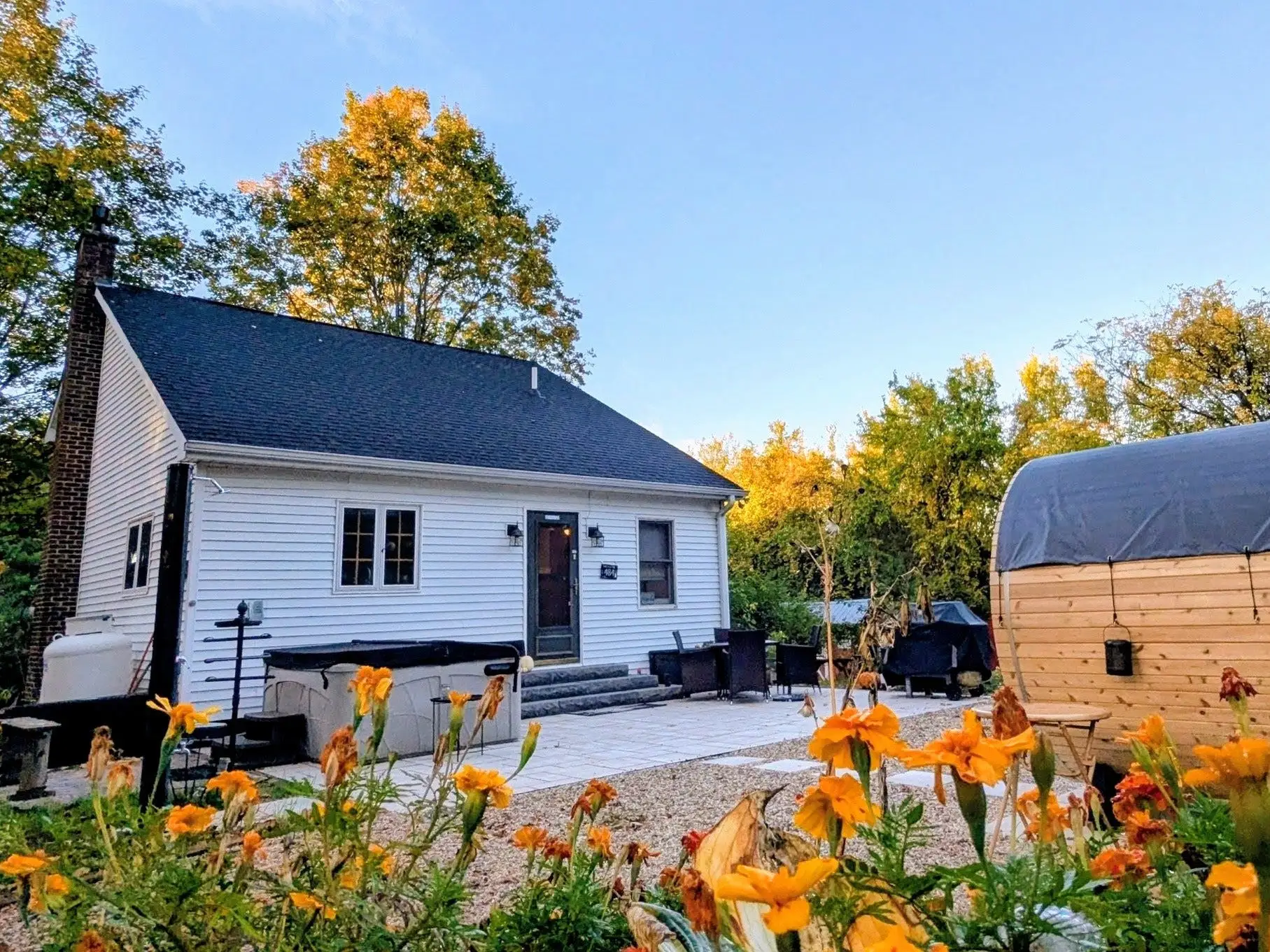 White house with patio, barrel sauna, orange flowers, and autumn trees under a clear blue sky.