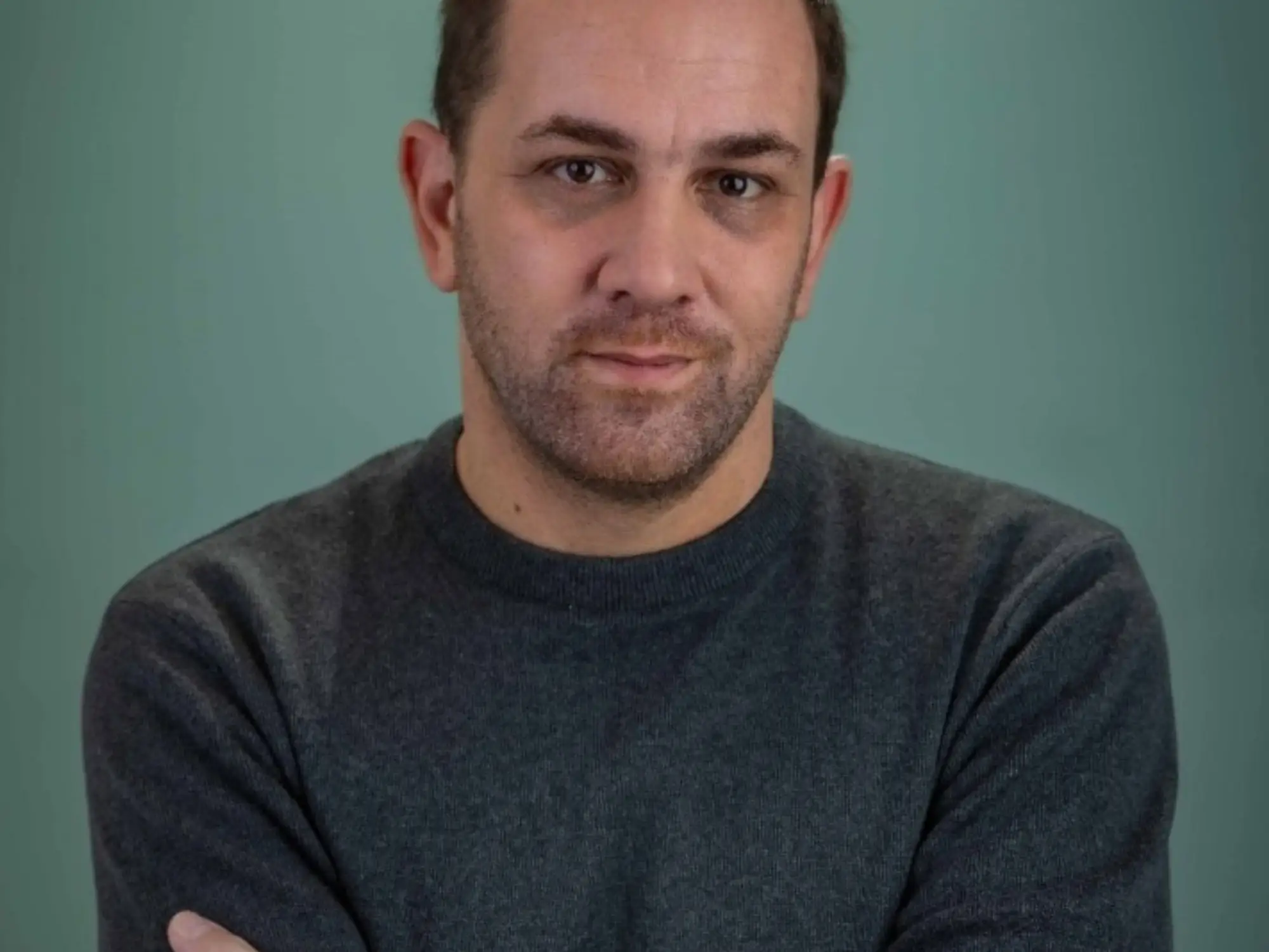 Portrait of a person in a dark sweater with arms crossed against a plain teal background.