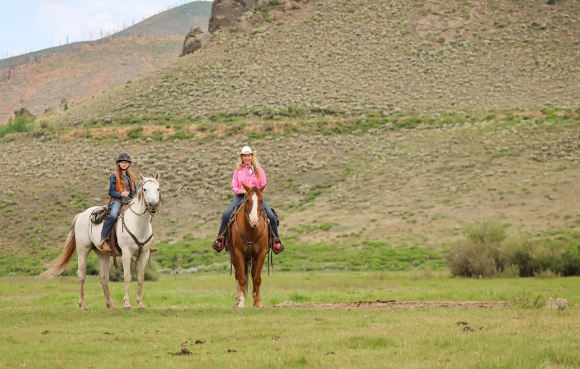 Two horseback riders pause in a grassy valley with rocky hills in the background.