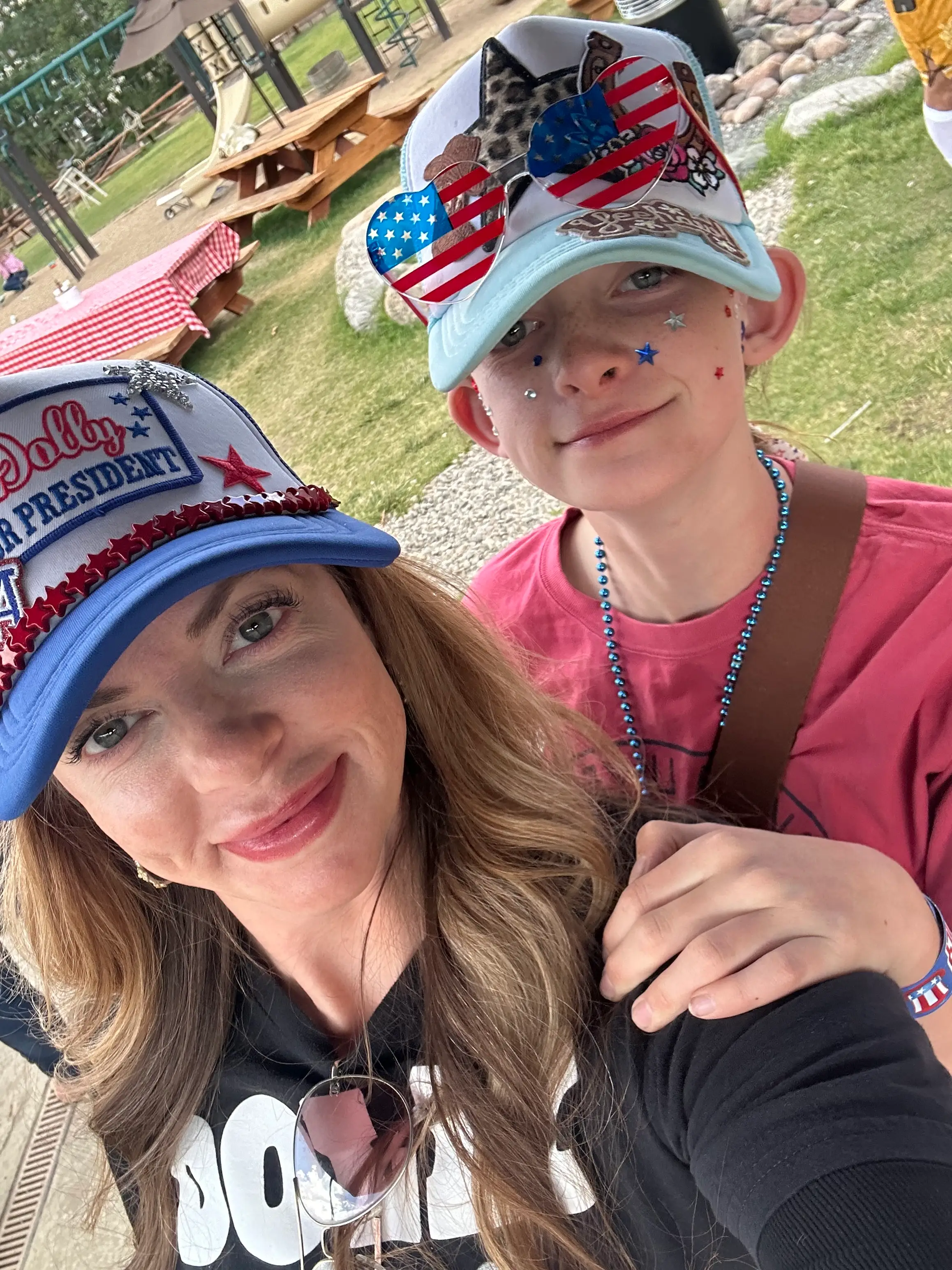 Two people wearing patriotic hats and star decorations pose for a selfie in an outdoor picnic area.