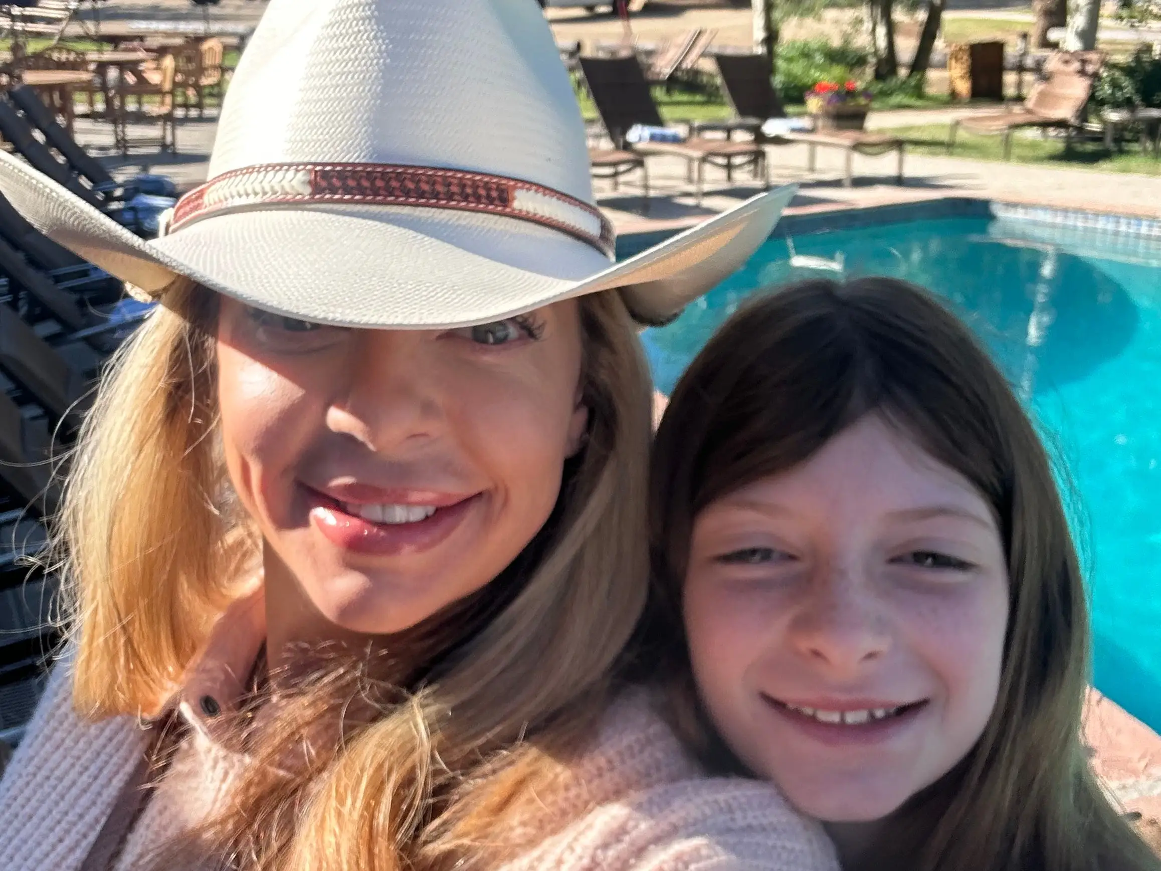 Two smiling people take a selfie beside an outdoor swimming pool with lounge chairs and trees in the background.
