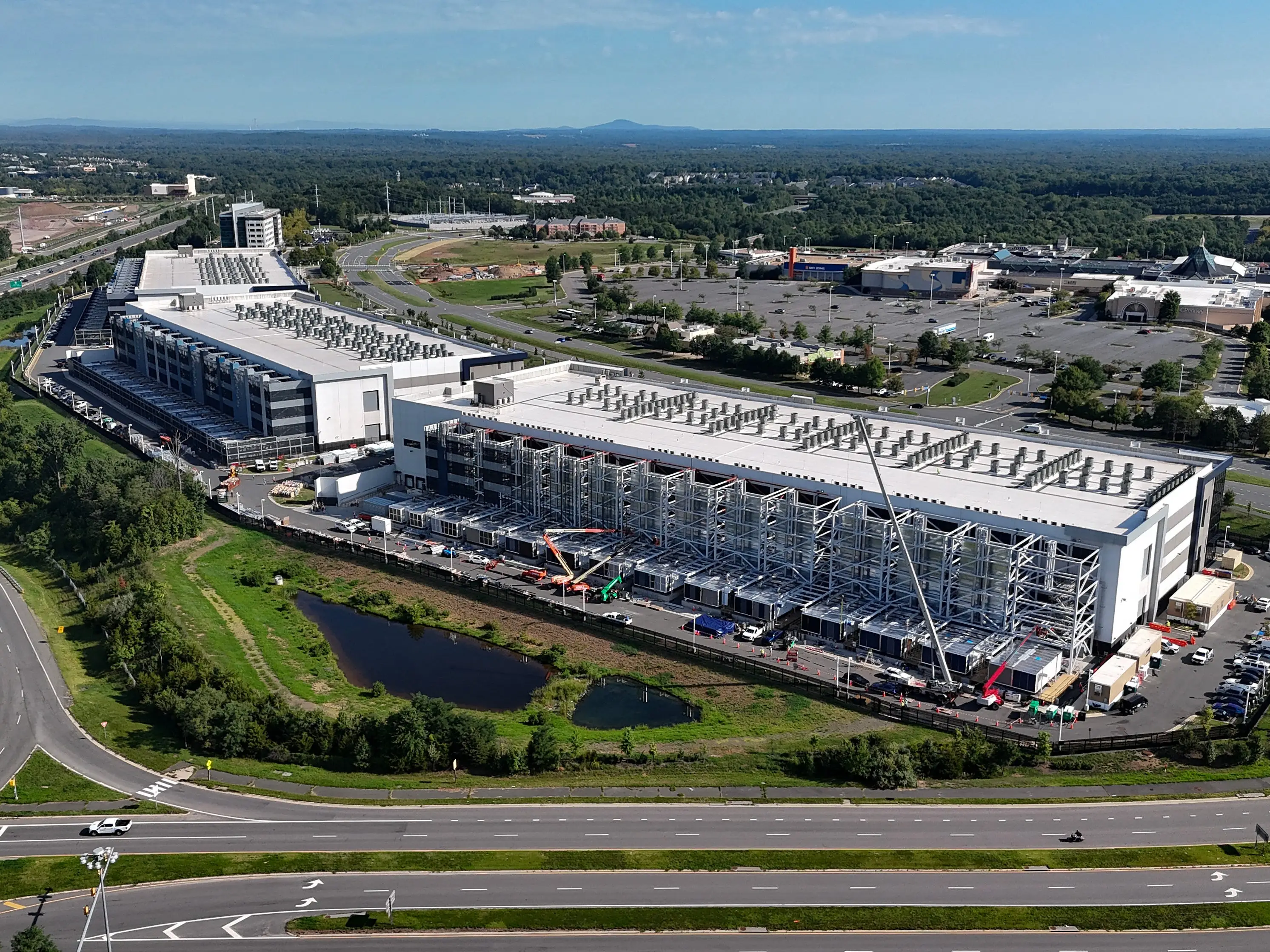 Aerial view of a large industrial facility with rooftop equipment, exterior construction, roads, parking lots, and surrounding greenery.