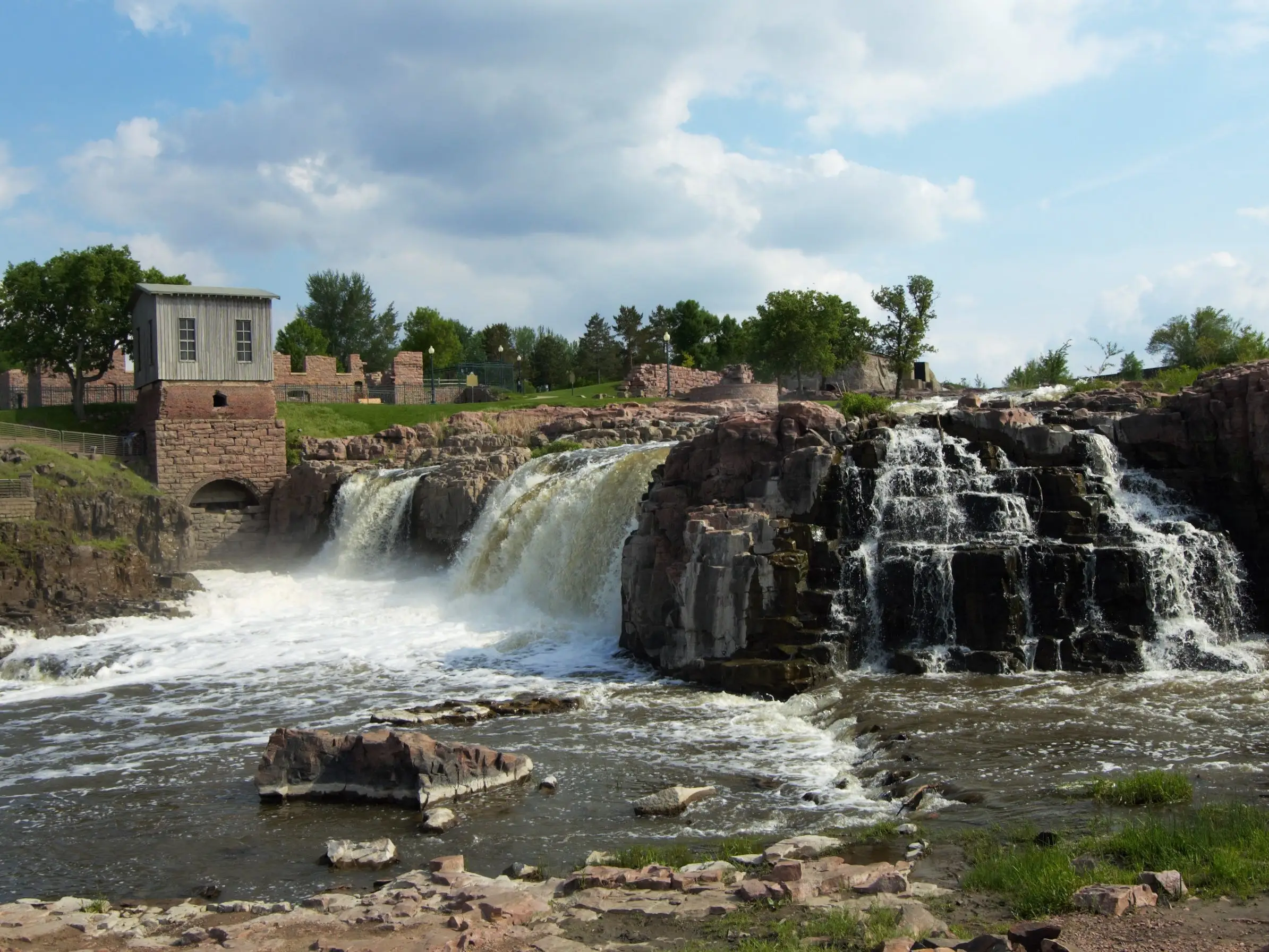 Waterfalls cascade over rocky ledges beside a stone-and-wood structure under a partly cloudy sky.