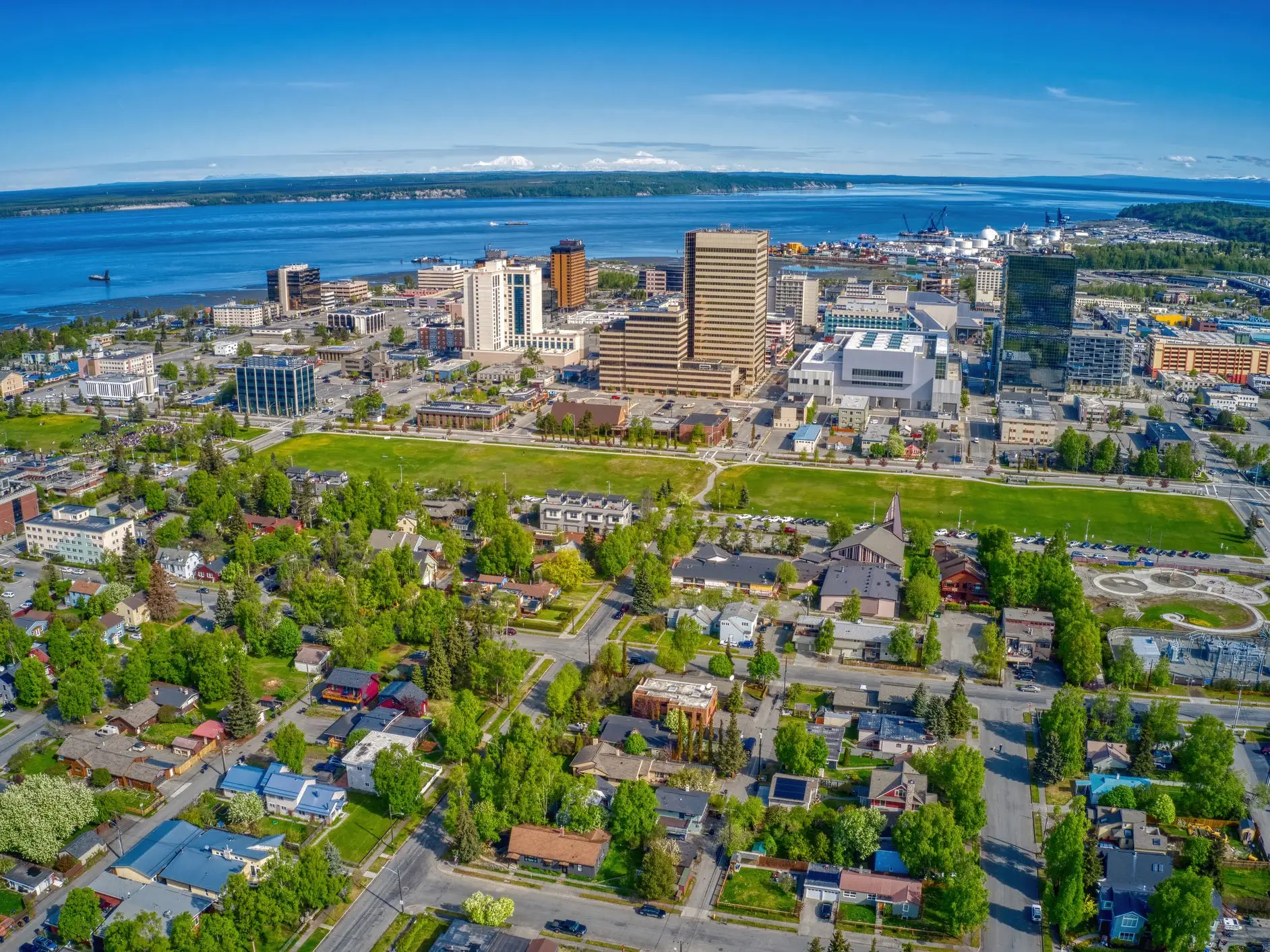 Aerial view of downtown Anchorage with residential neighborhoods, high-rise buildings, green parkland, and Cook Inlet waterfront.