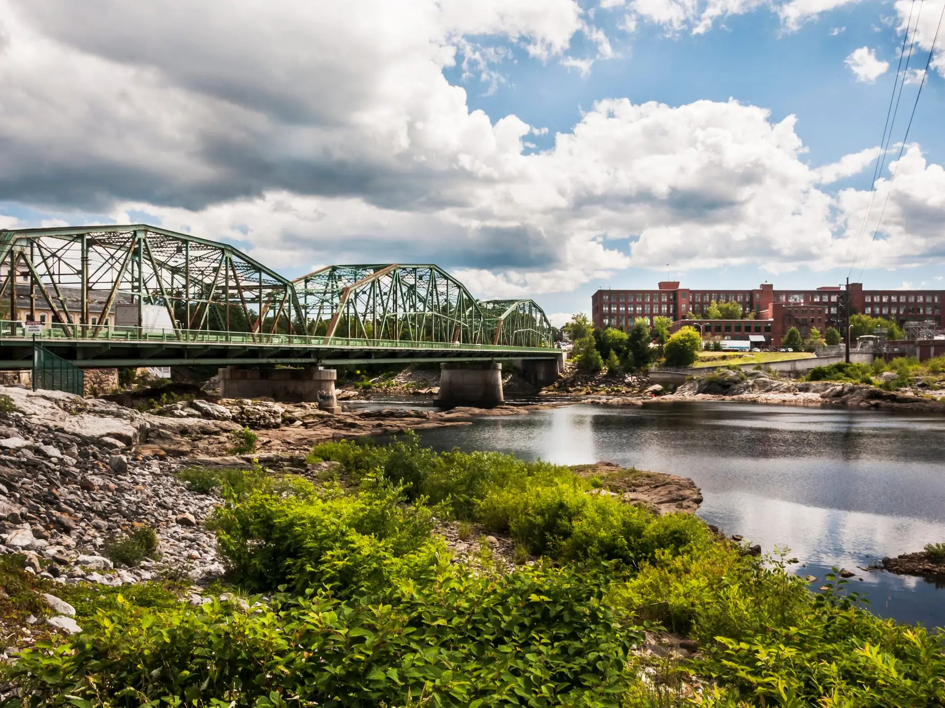 Green steel truss bridge spans a calm river beside rocky banks, vegetation, and red brick industrial buildings.