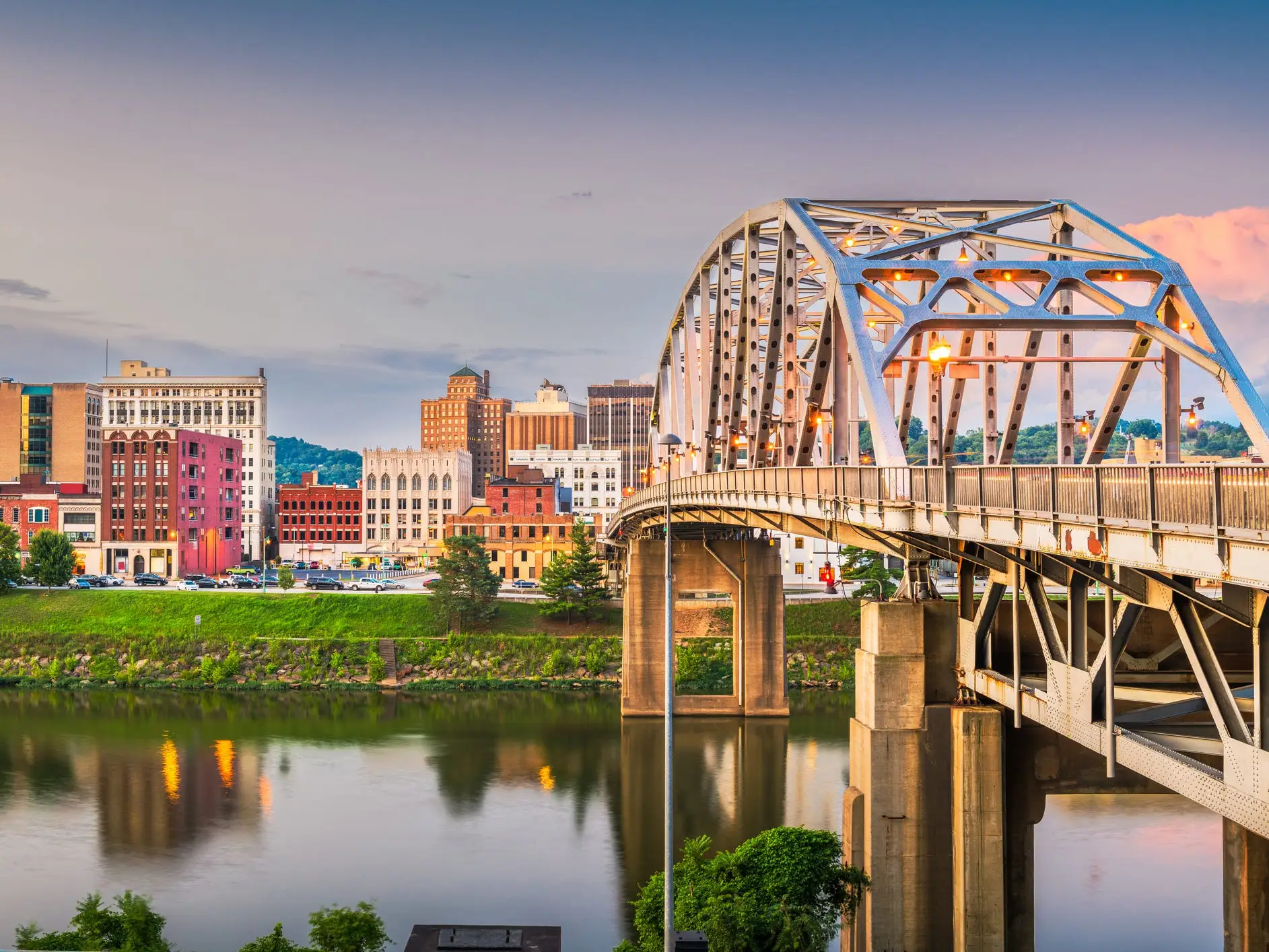 Steel arch bridge spans a river beside a downtown skyline at dusk with reflections on the water.