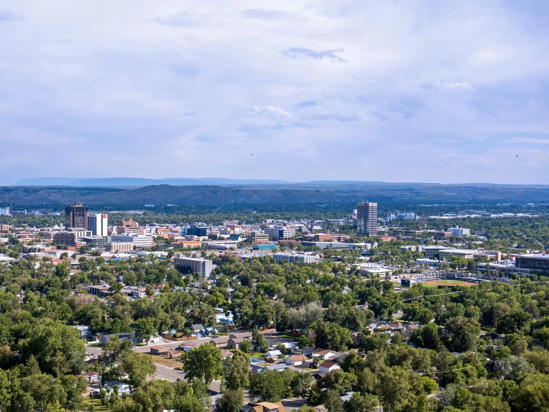 Elevated view of a city skyline with tree-filled neighborhoods and low hills under a cloudy sky.