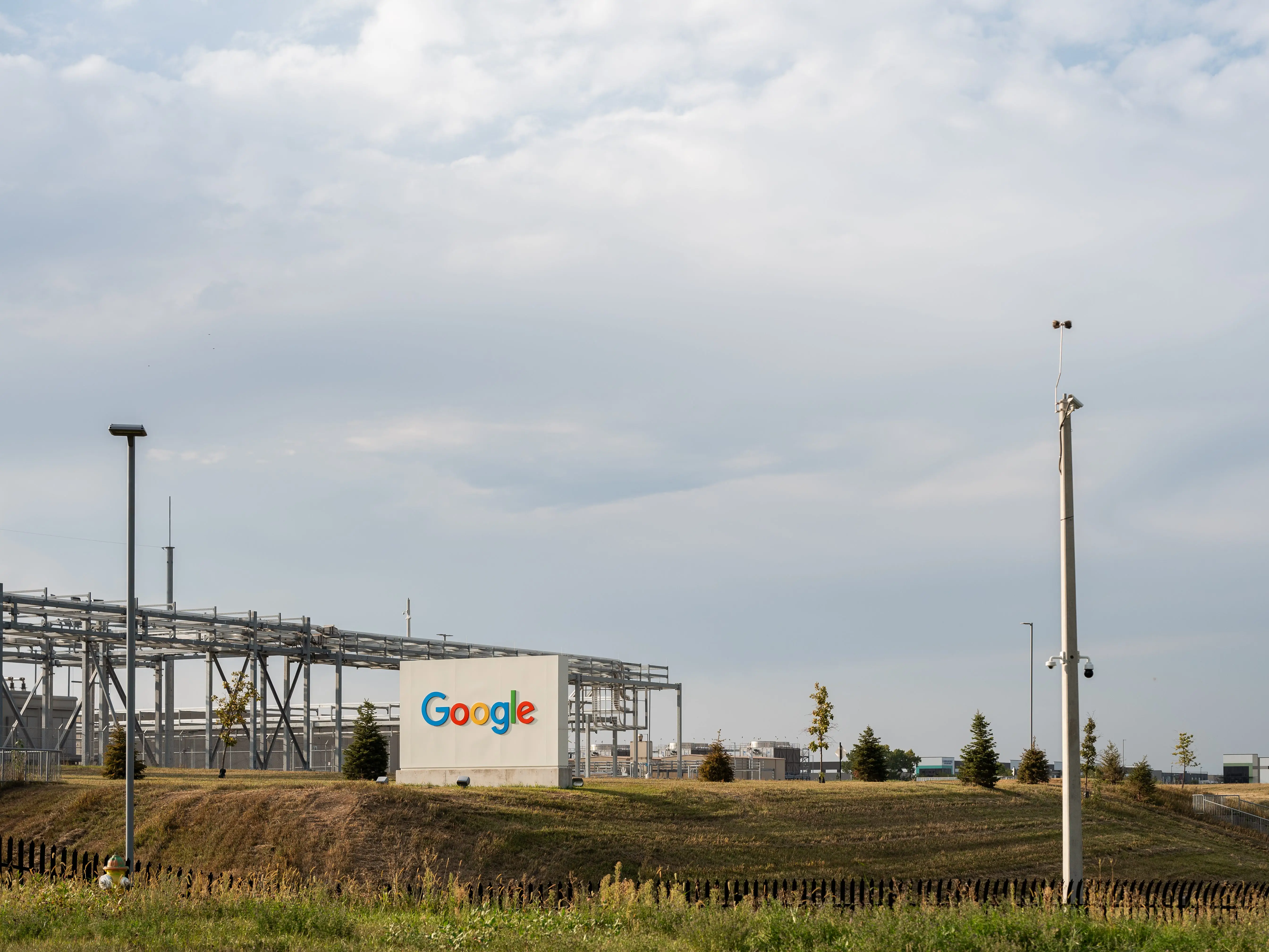 Google logo appears on a white industrial building beside utility structures, poles, grass and a cloudy sky.