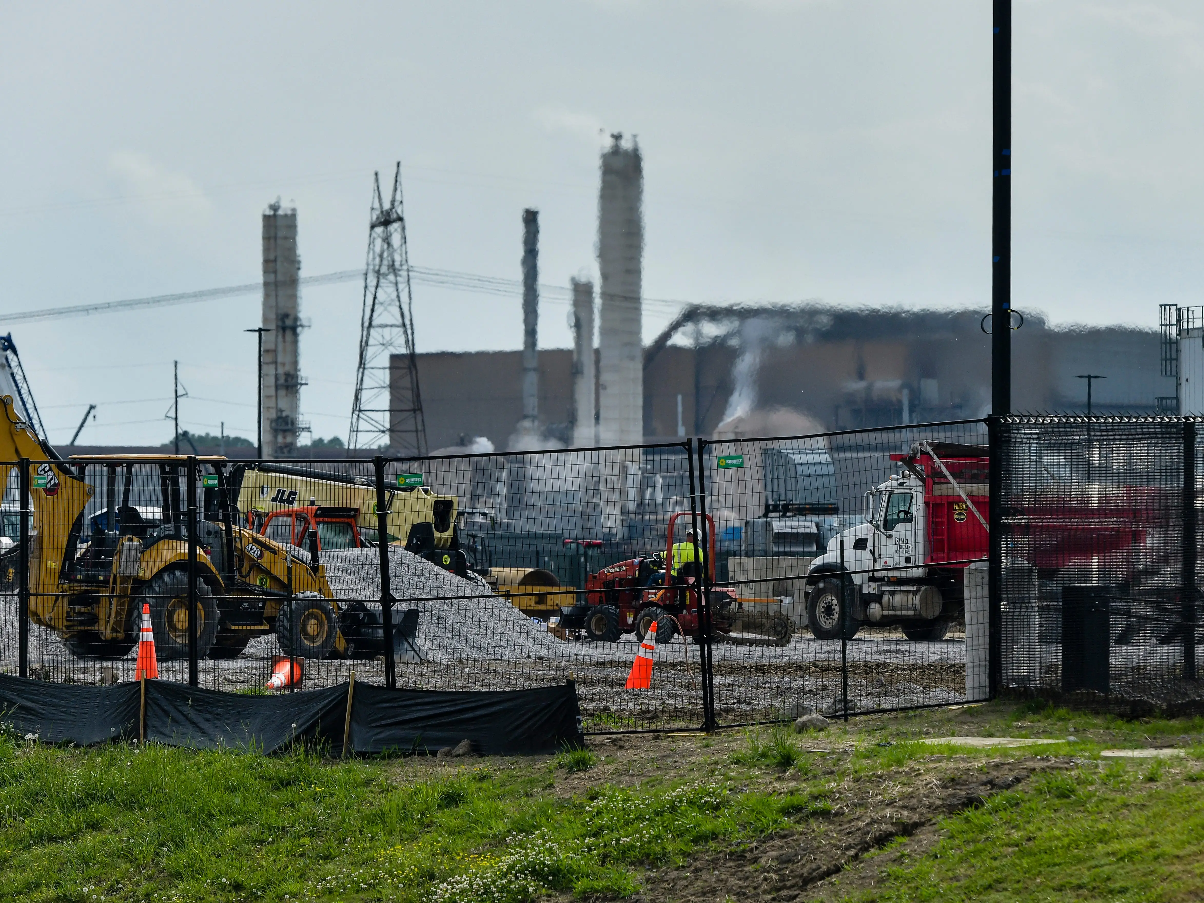 Industrial construction site behind a chain-link fence with heavy equipment, trucks, tall towers, and drifting smoke.