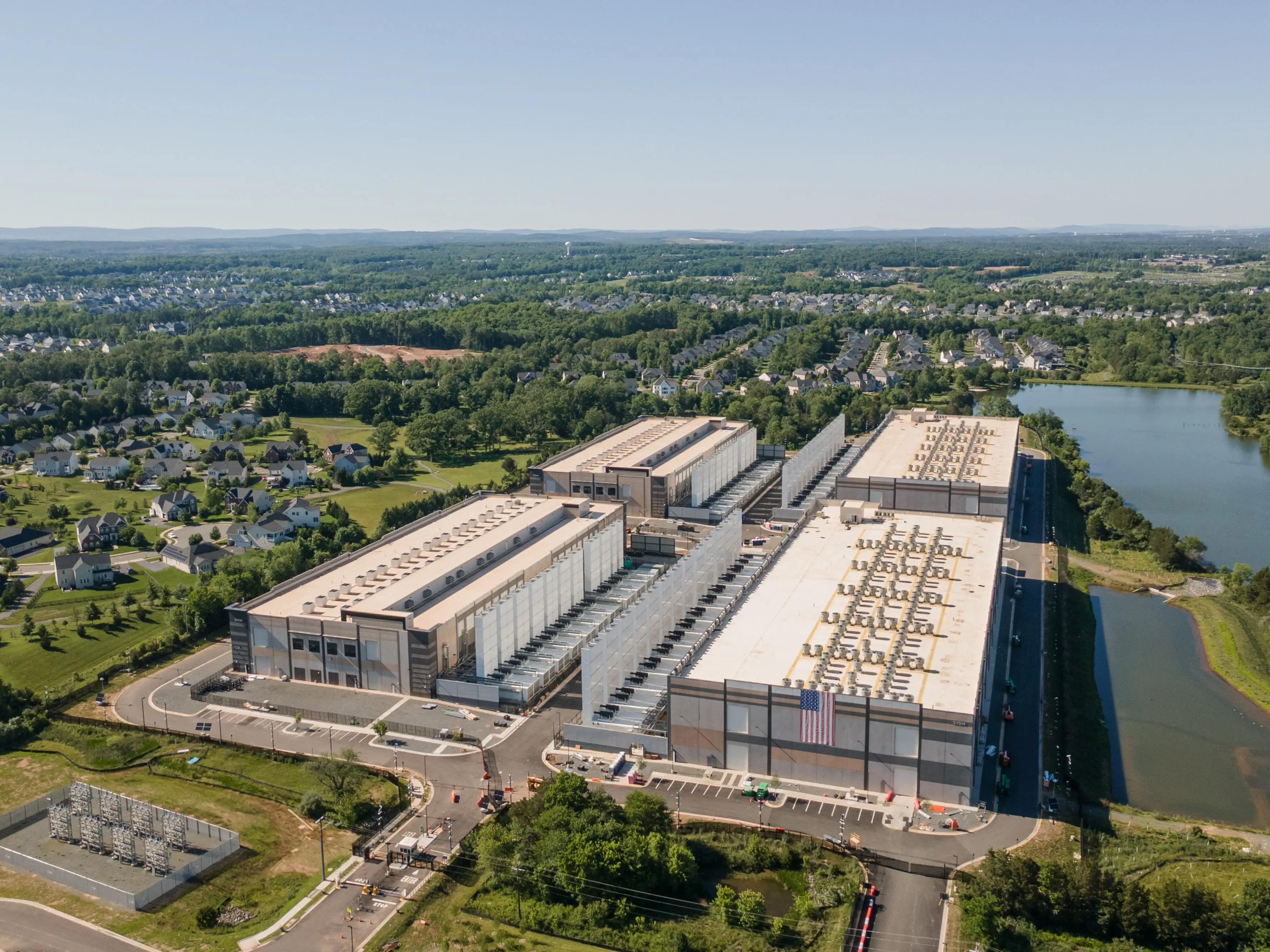 Aerial view of large industrial data center buildings beside a river and surrounding suburban neighborhoods.