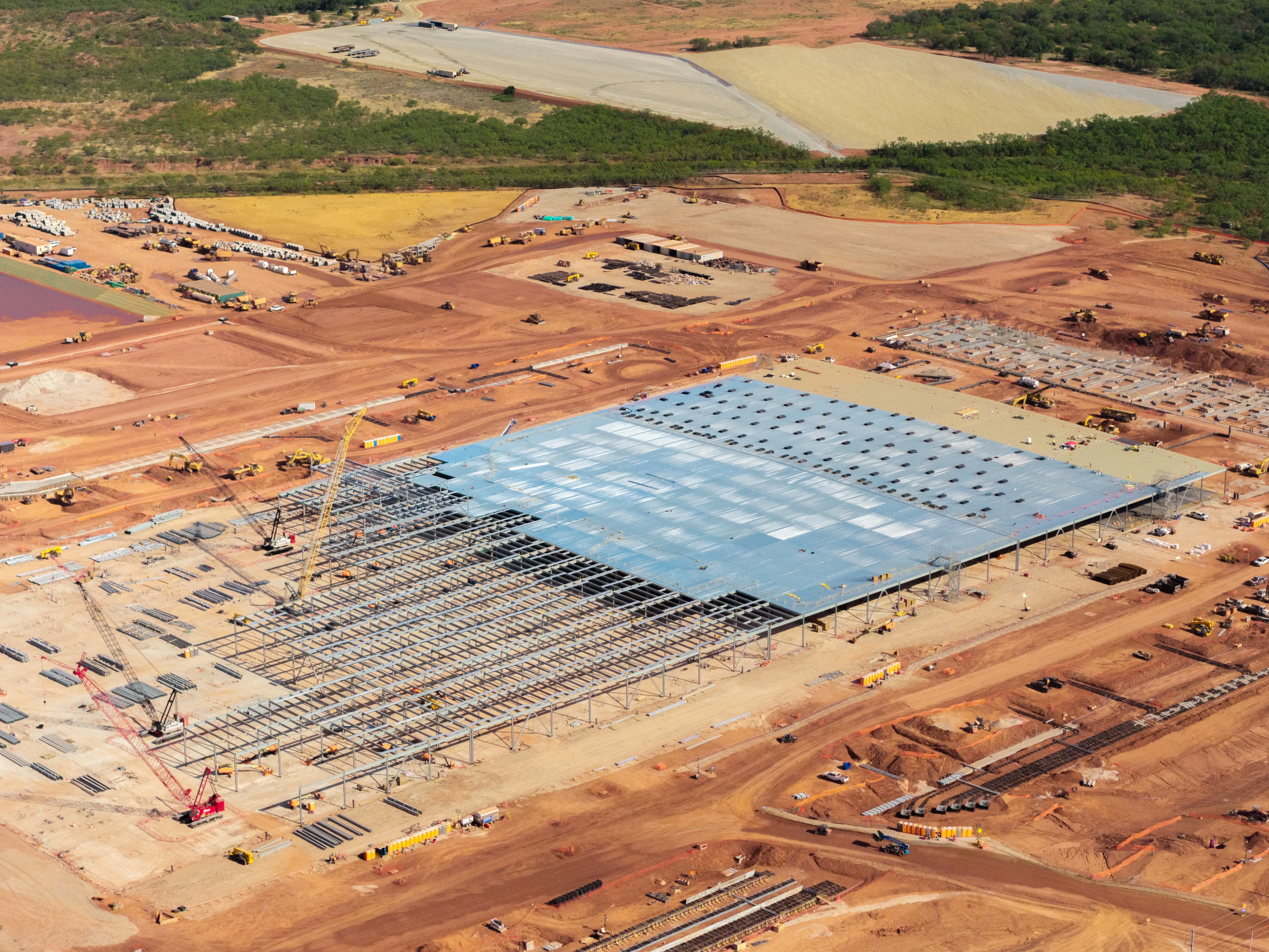 Aerial view of a large industrial building under construction amid red dirt earthworks, cranes, equipment, and surrounding forest.