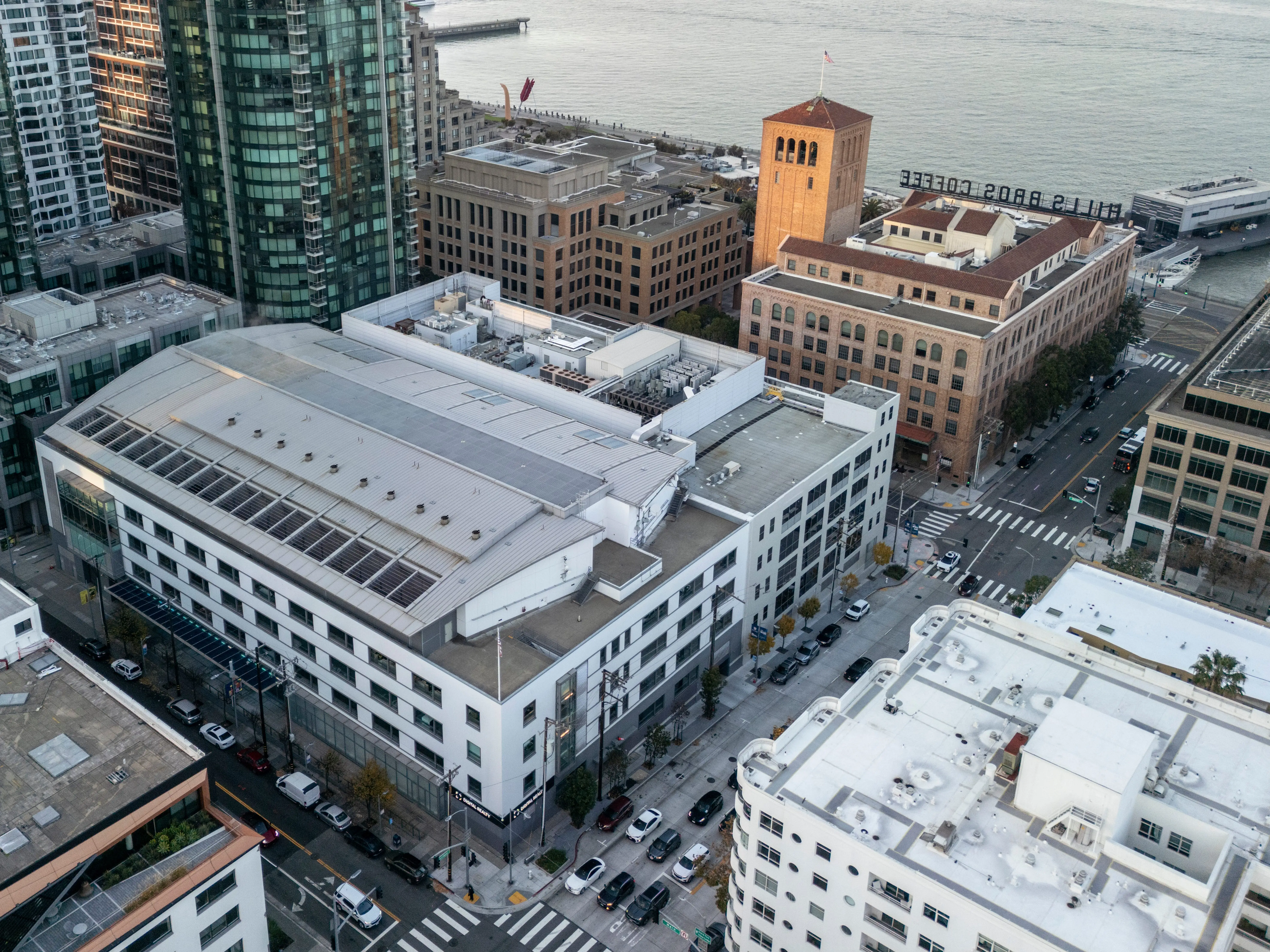Aerial view of city blocks with modern buildings, streets, cars, and a waterfront pier beside the bay.