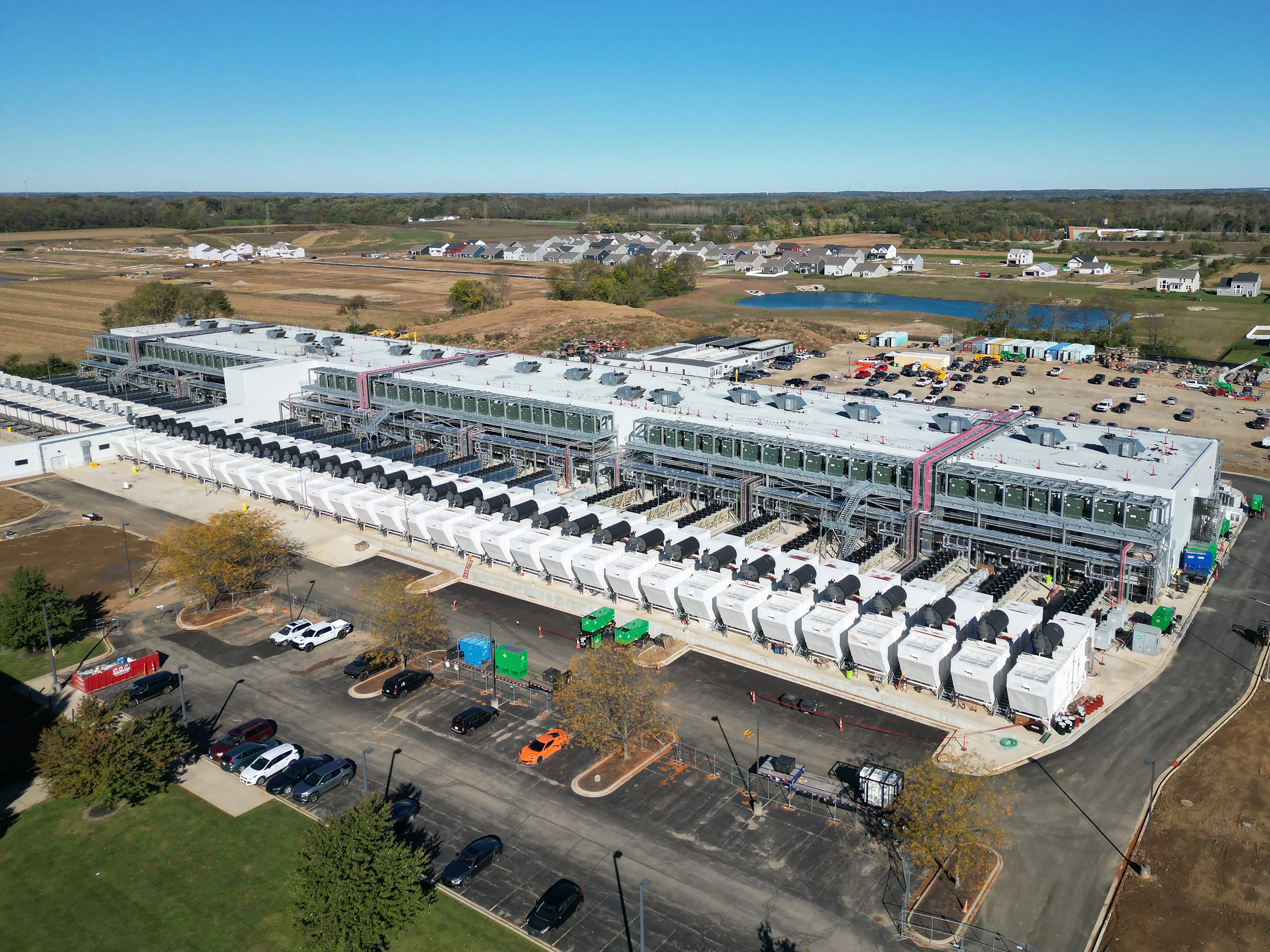 Aerial view of a large industrial construction site with rooftop equipment, rows of white units, cars, and surrounding fields.