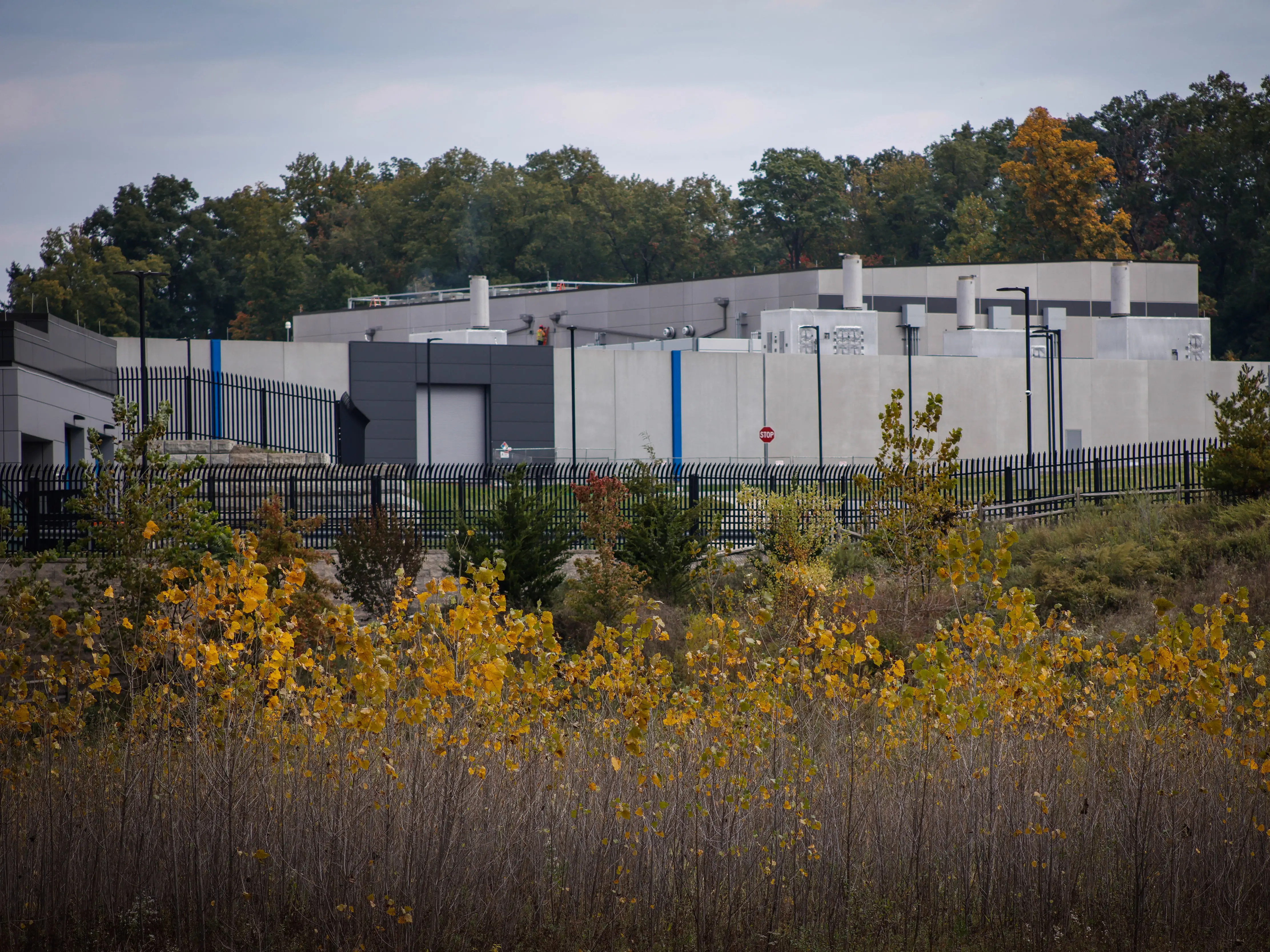 Gray industrial facility behind black security fencing, with autumn vegetation and trees under an overcast sky.